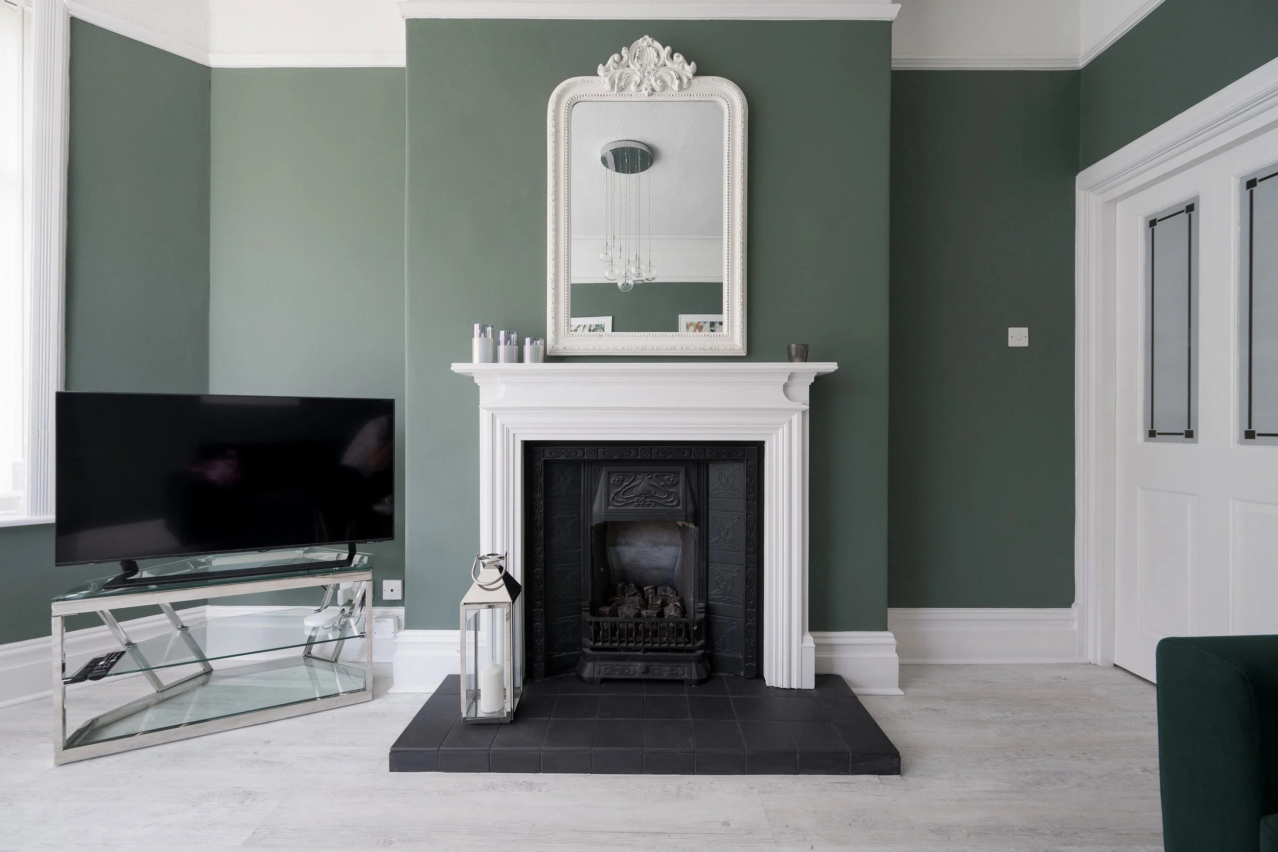 Living room with green walls, white crown molding, a decorative white fireplace mantel, a large mirror above the fireplace, and a black fireplace insert. To the left is a sleek glass TV stand with a flat screen TV and remote controls. Candles and can
