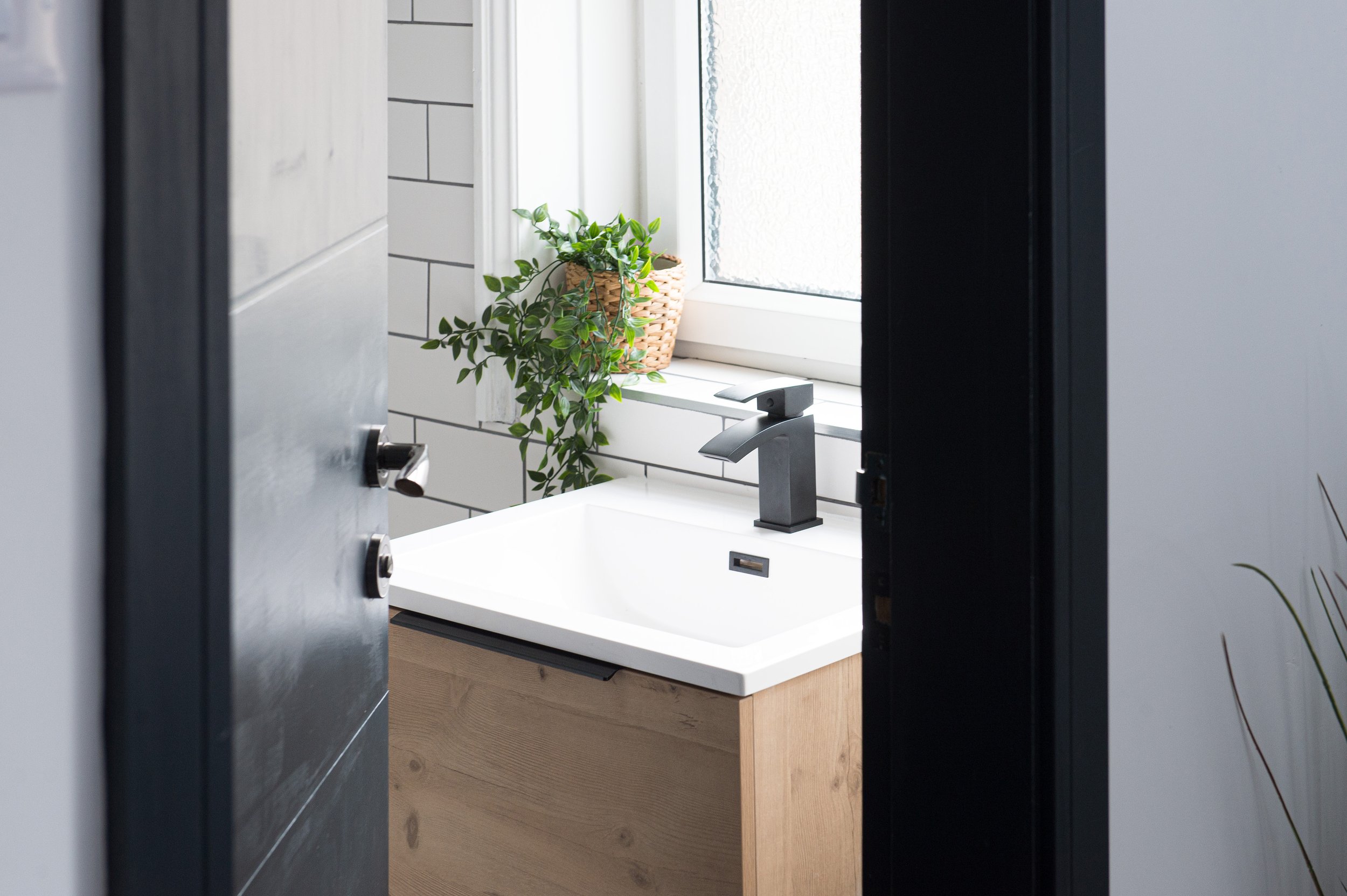 The image shows a modern bathroom with a partial view of a open door. Inside, there is a white sink with a silver faucet, a window with frosted glass, and a potted green plant in a woven basket on the window sill. The bathroom has white tiled walls w