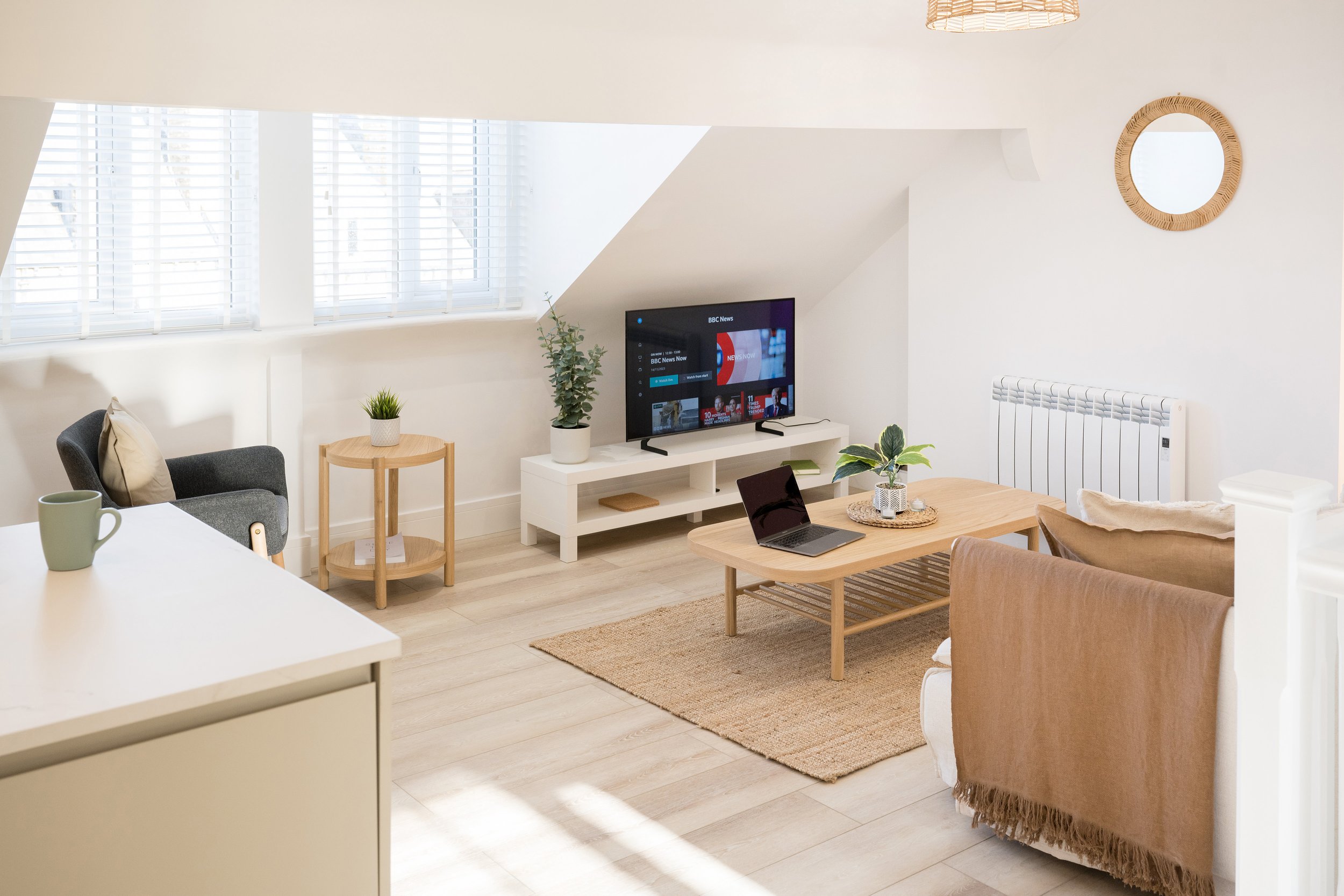 Bright living room with white walls and large windows with white blinds. Contains a gray armchair with a beige pillow, a round wooden side table with a small plant, a white TV stand with a TV showing BBC News, a laptop on a wooden coffee table, a bei