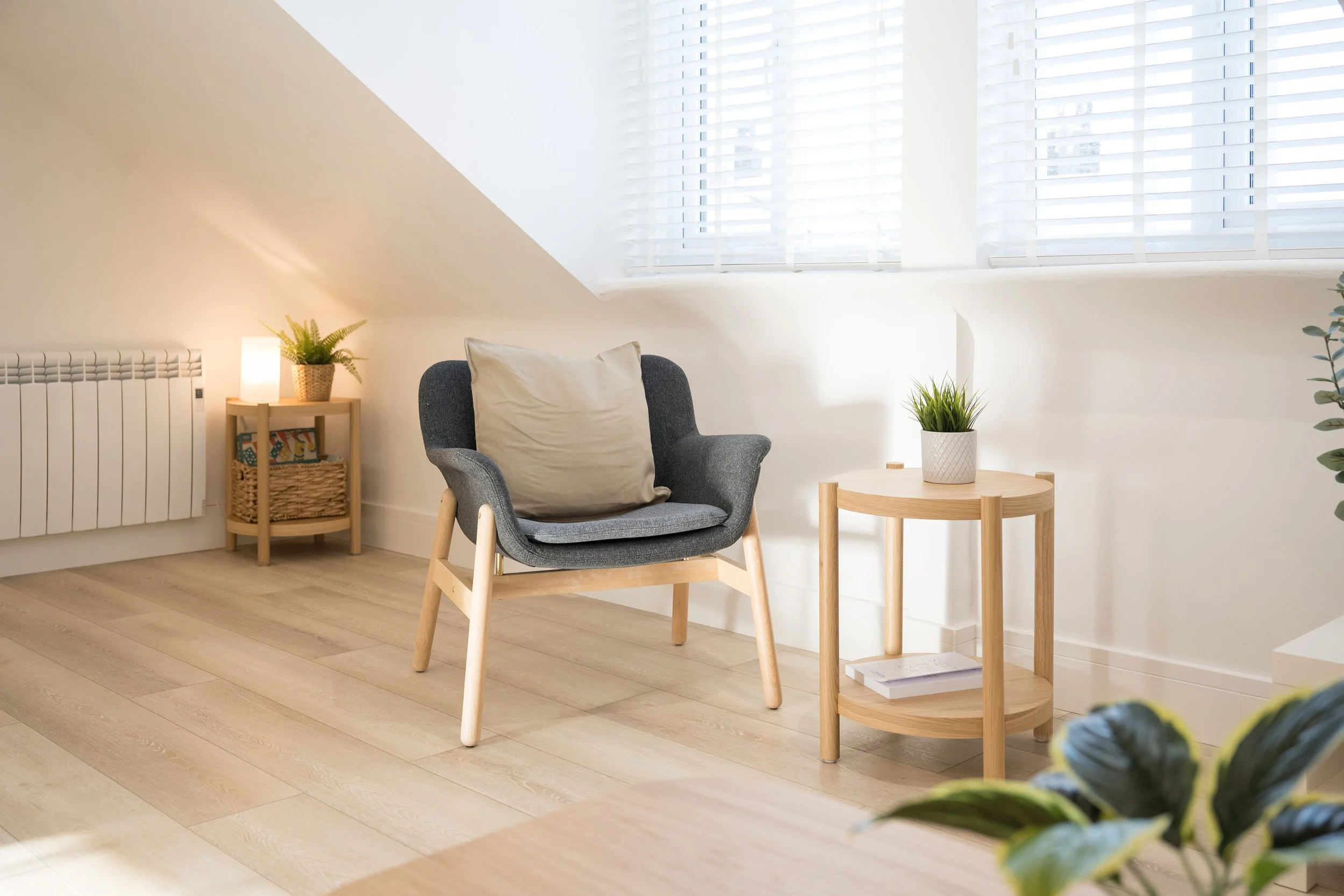 A cozy corner of a living room with a gray armchair, a light wooden side table with a potted plant, and another small round wooden table with a white pot of greenery. Natural light filters through white blinds on a large window, creating a bright and