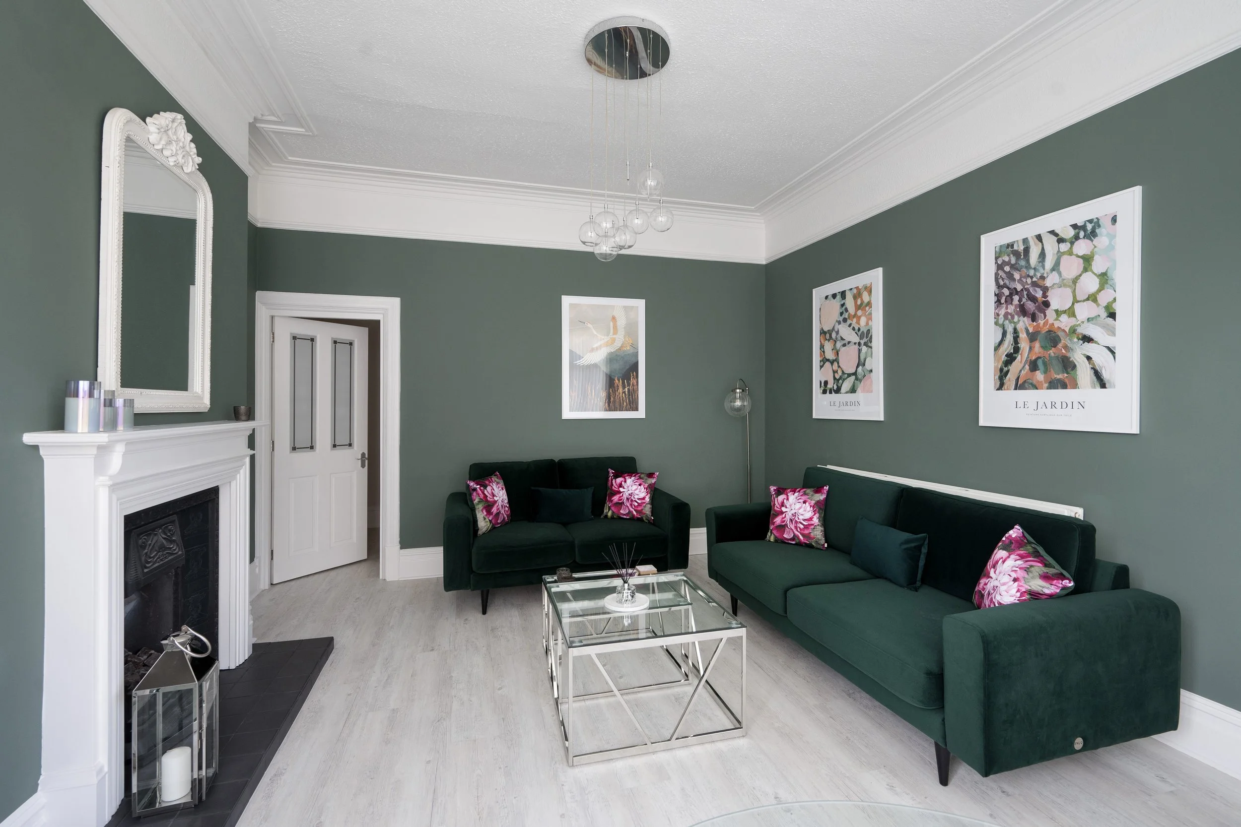 Living room with green walls, white ceiling moldings, a white fireplace, and framed artwork. Two green sofas with pink floral cushions, a glass coffee table, and a modern chandelier with hanging glass globes.