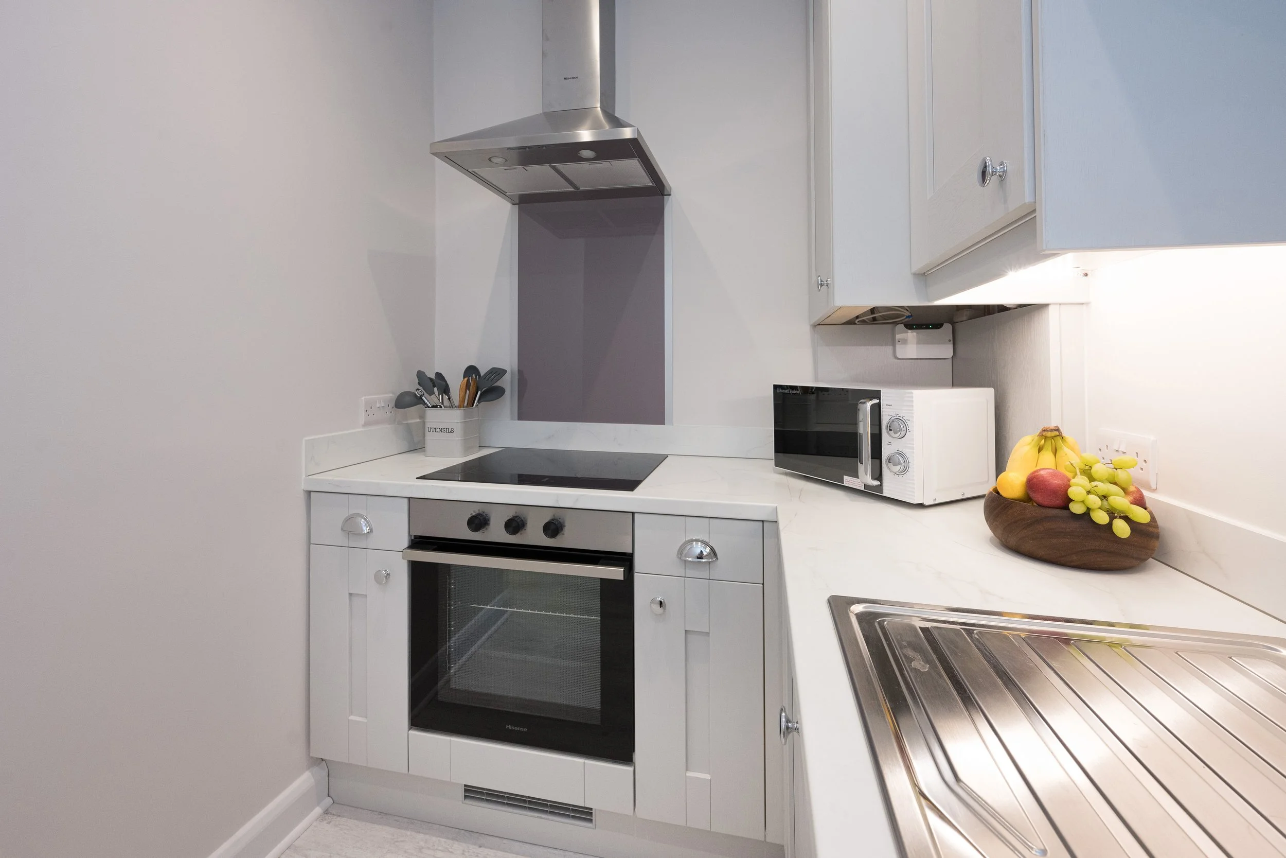 A modern kitchen with white cabinets, a black oven, a microwave, a metal range hood, and a countertop with a fruit bowl containing bananas, mangoes, and grapes.