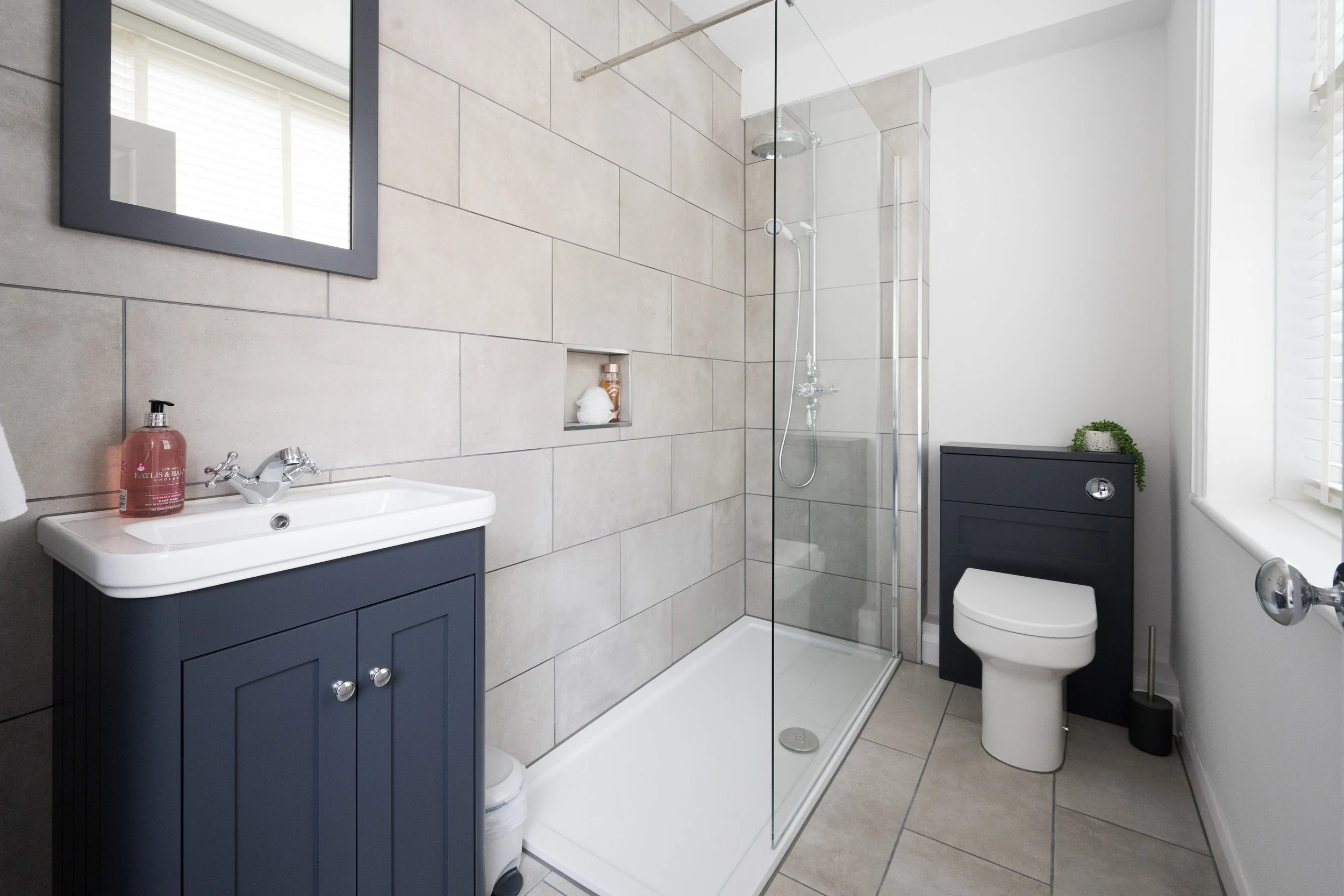 Modern bathroom with beige tiled walls, a glass-enclosed shower, navy blue vanity with silver knobs, white toilet, small green plant, and a mirror with a black frame.