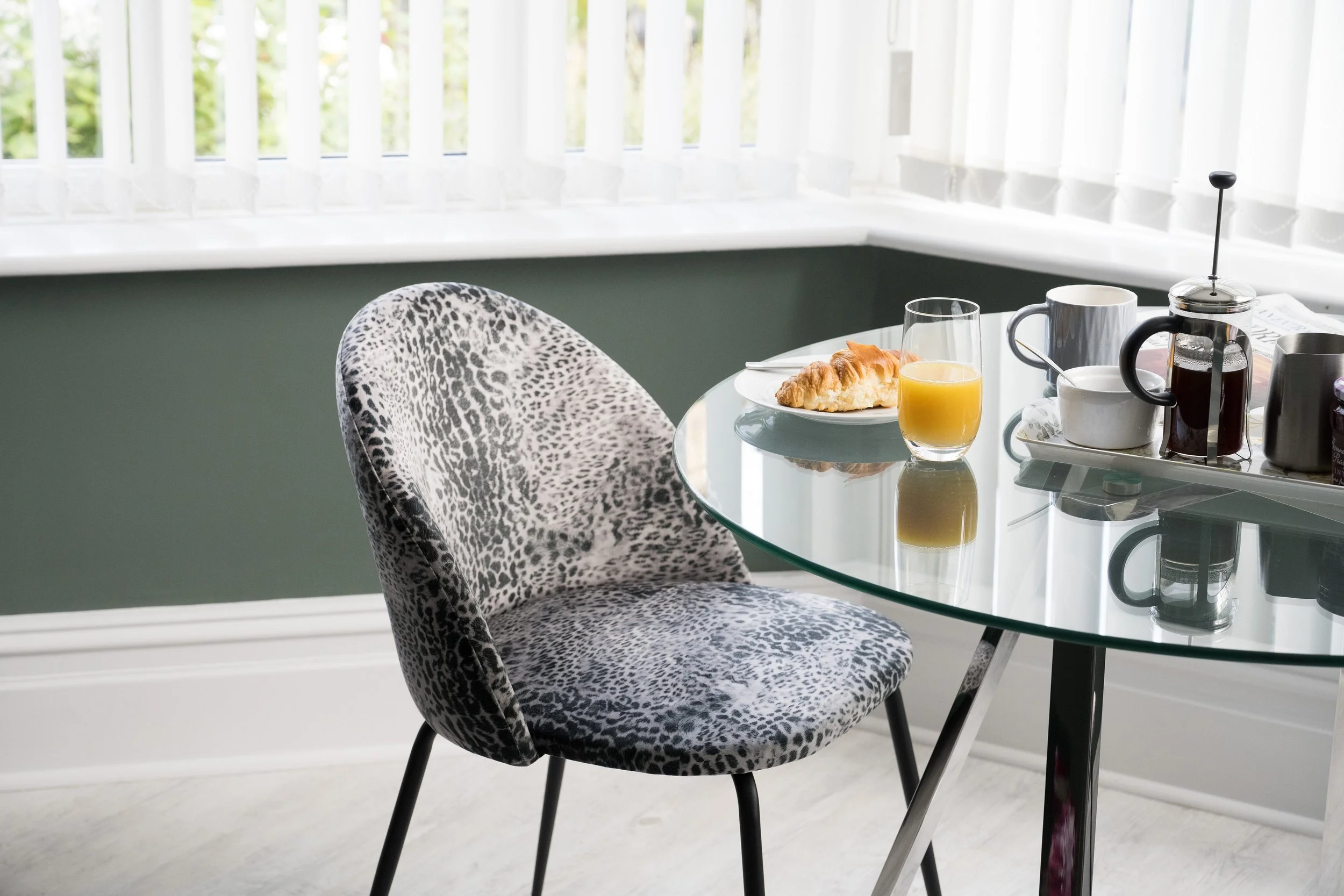 A glass dining table with a plate of croissants, a glass of orange juice, a mug, a French press coffee maker, and a coffee mug. A leopard print upholstered chair is partially visible in the foreground, and there is a window with white blinds in the b