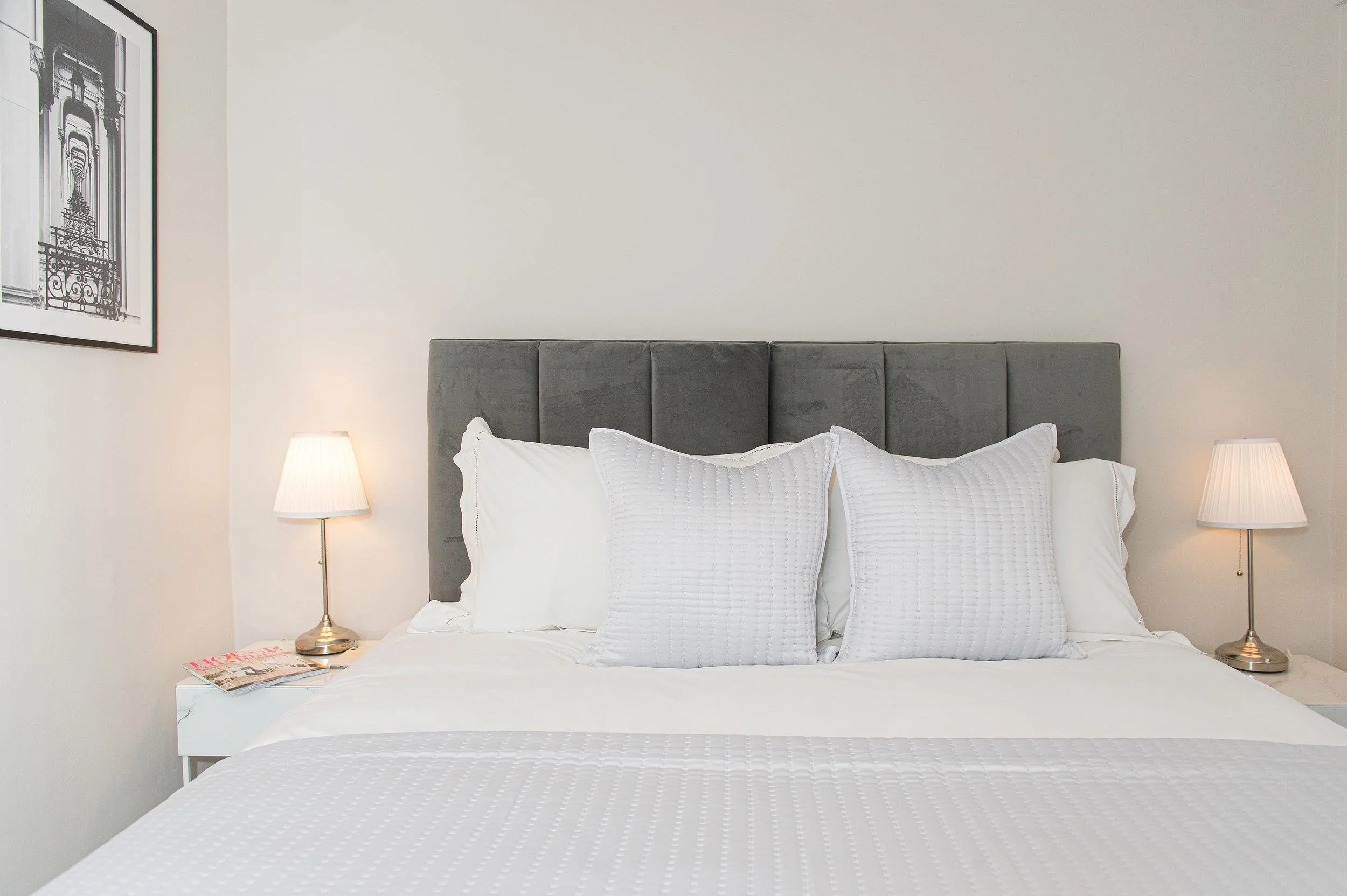 A bedroom with a grey upholstered headboard, white bedding, and pillows. There are two matching bedside lamps on either side of the bed, and a framed black-and-white architectural print on the wall to the left.