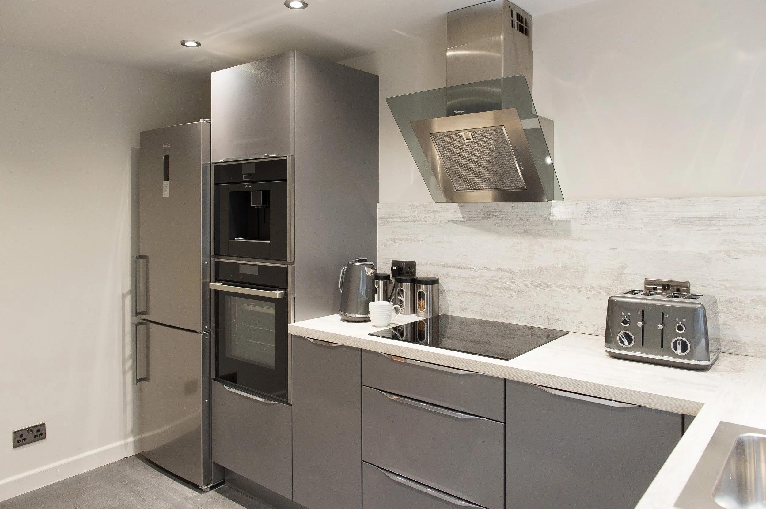 Modern kitchen with stainless steel refrigerator, oven, and range hood, white countertop with coffee mugs and appliances, and gray cabinetry.