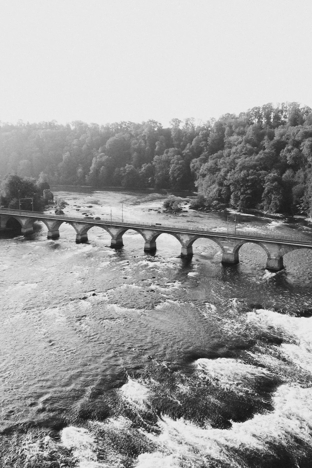 Black and white photo of a river with a stone arch bridge crossing over it, surrounded by trees.
