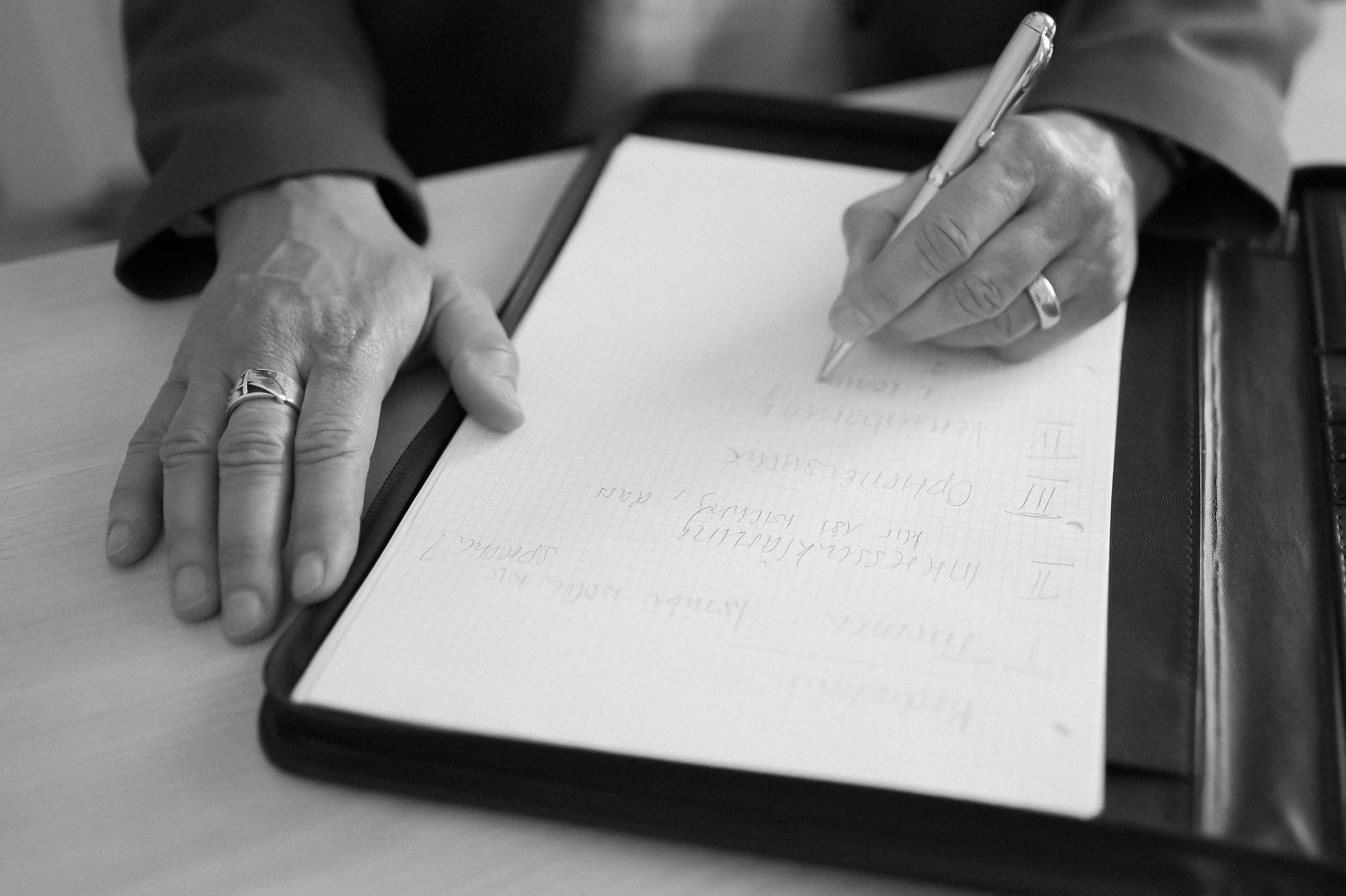 A person writing notes in a notebook with a pen, wearing a ring on their finger, and a dark long-sleeve shirt. The notebook is on a flat surface, and the person's hands are visible.