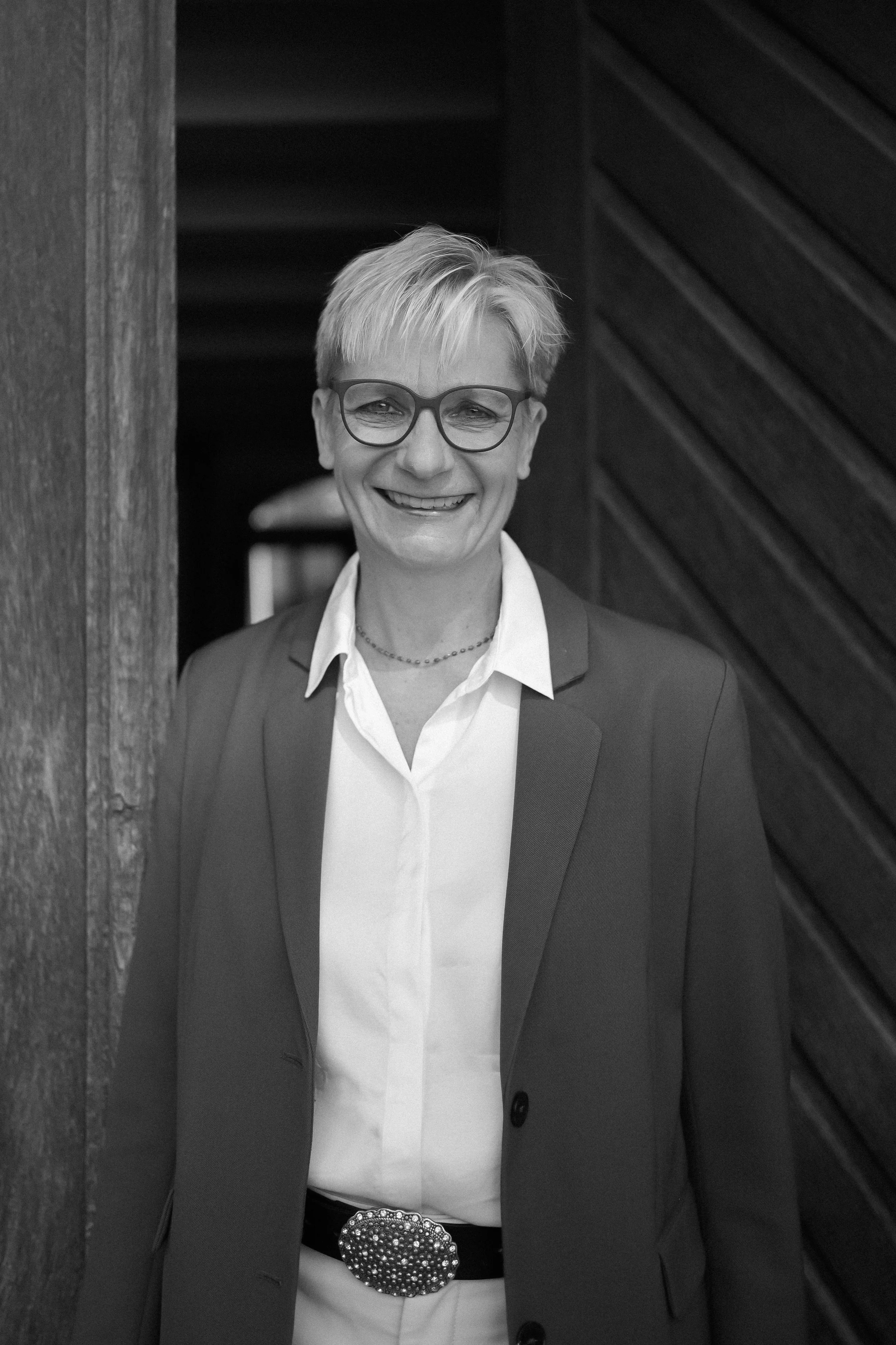 A woman with short blond hair and glasses smiling, wearing a suit with a white shirt and a belt with a large, gemstone-studded buckle, standing in front of a wooden wall.