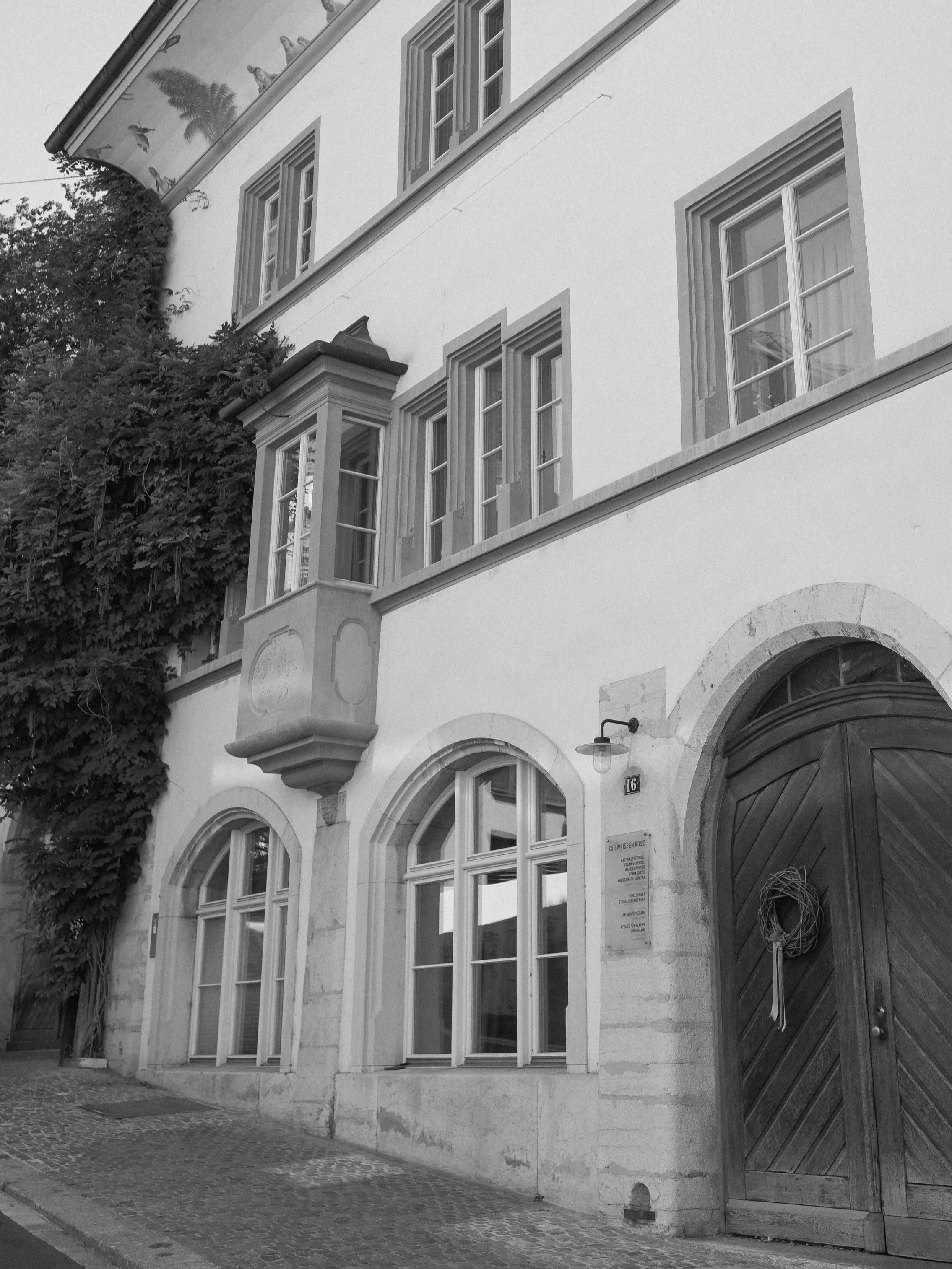Black and white photo of a building with arched windows and a large wooden door. There is a street lamp next to the door and dense shrubbery on the left side of the building. The building has a decorative corner element above the door and a sign below the street lamp with text.