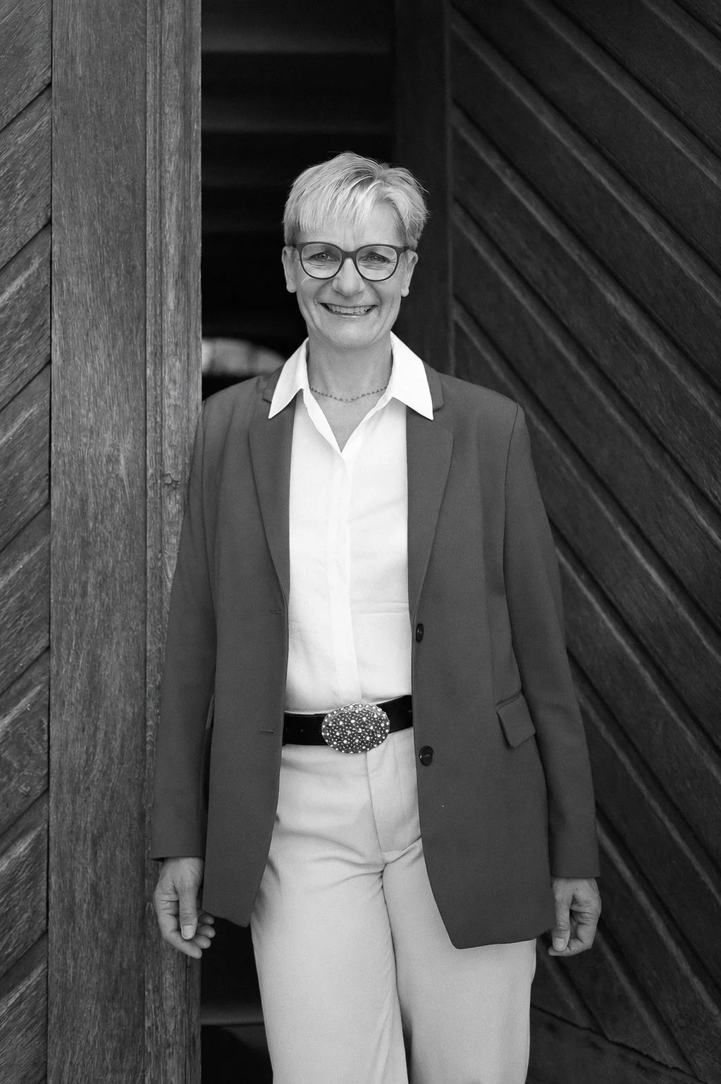 A woman with short hair and glasses smiling, dressed in a suit with a white shirt, standing in front of a wooden wall.