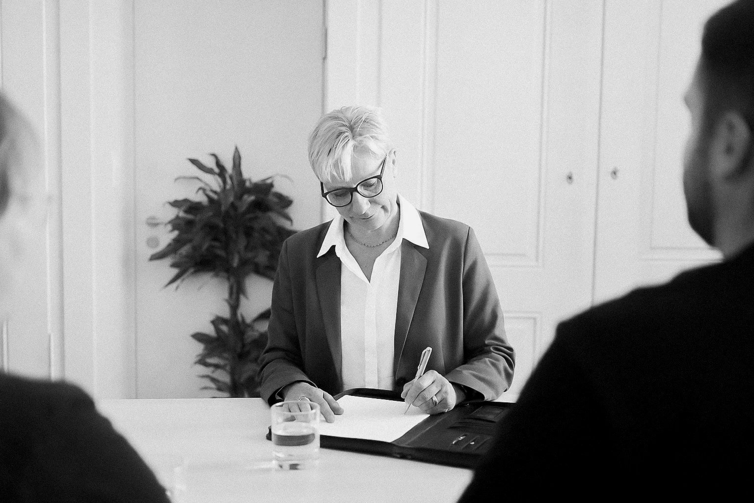 A woman with short light hair, glasses, and a blazer is sitting at a table, writing on a notepad with a pen. In front of her is a glass of water, and she is surrounded by two other people whose shoulders are visible in the foreground. The background shows closed cabinet doors and a potted plant.