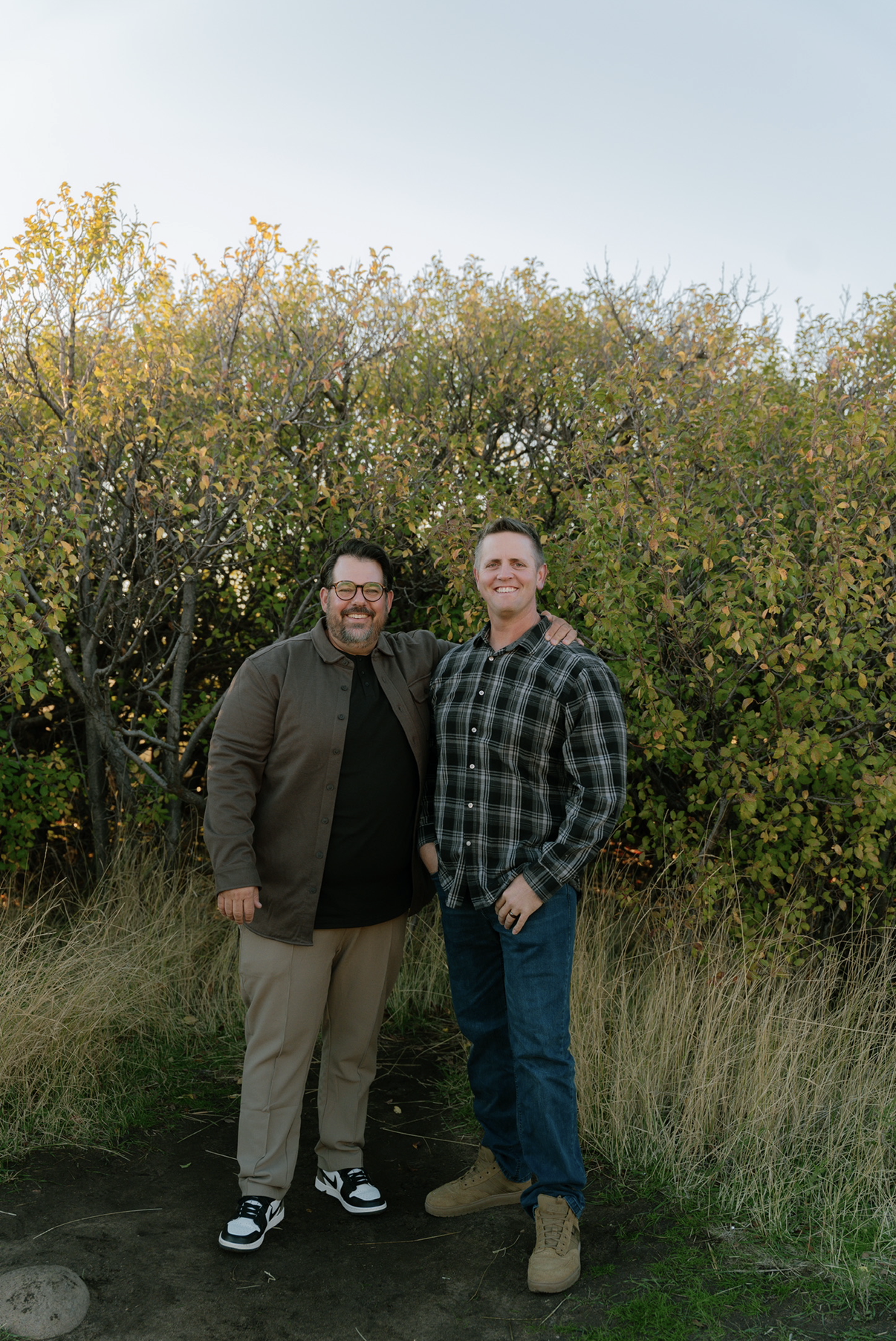 Two men standing outdoors in front of a bush with green leaves, smiling, with grass and dirt on the ground.