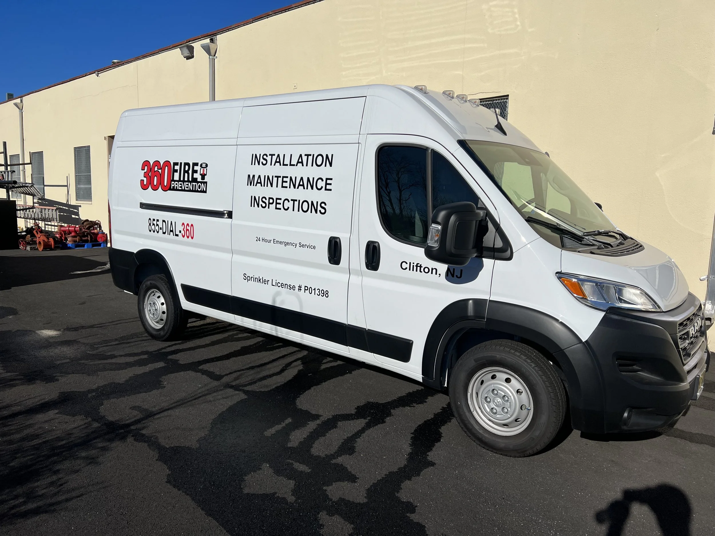 A white service van with black and red text advertising installation, maintenance, and inspections services. The van is parked outside a building with a beige wall and a small window, under a clear blue sky.