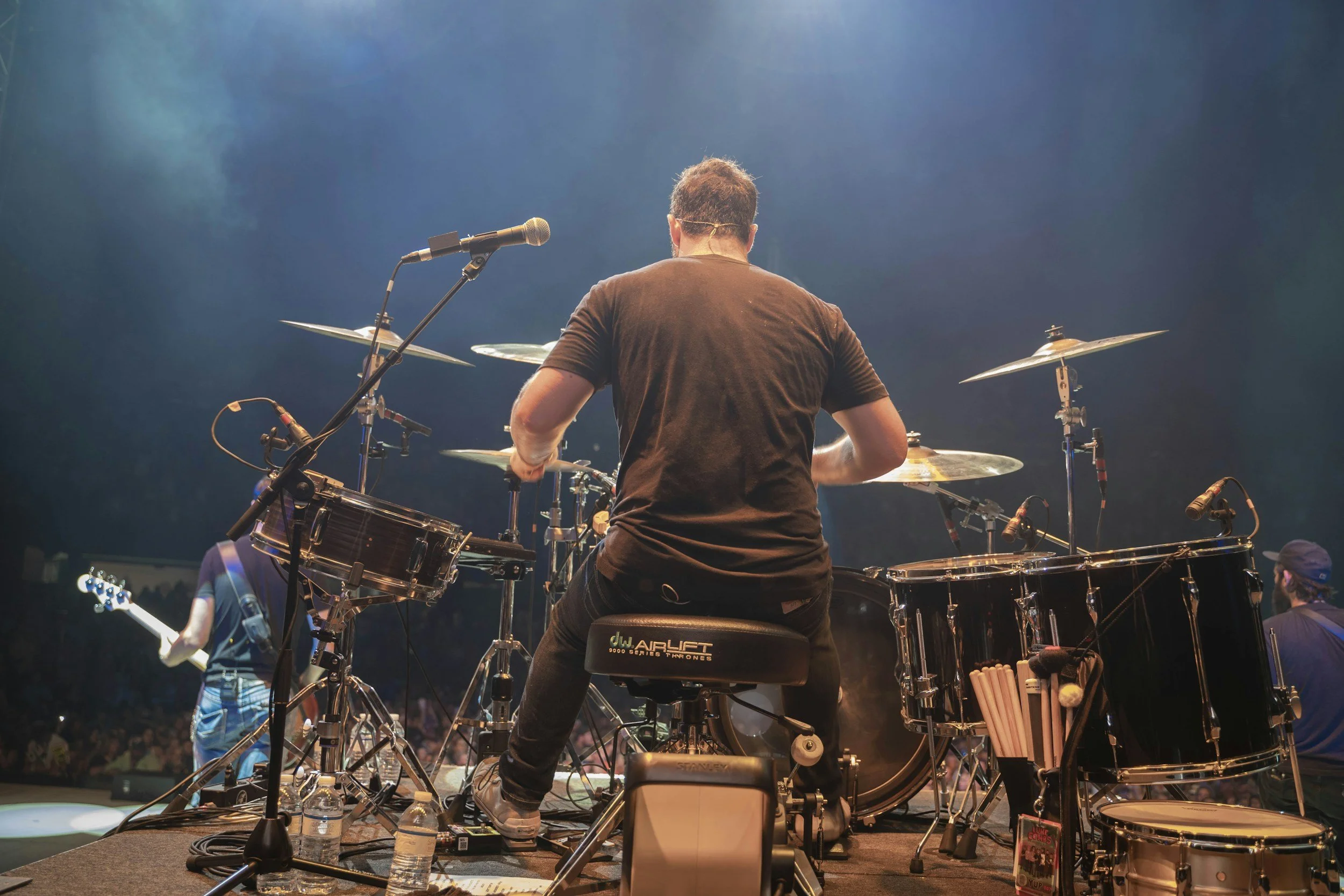 A musician playing drums on stage, seen from behind, with various percussion instruments and music equipment, stage lights illuminating the scene, and multiple band members visible in the background.