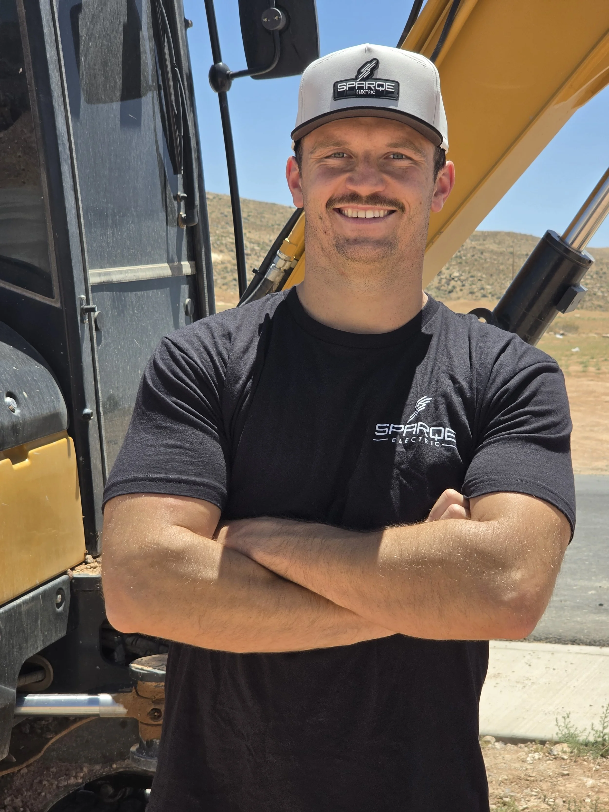 Smiling man with crossed arms wearing a gray cap with 'Sparqe Electric' logo and a black t-shirt with the same logo, standing in front of construction equipment outdoors on a sunny day.