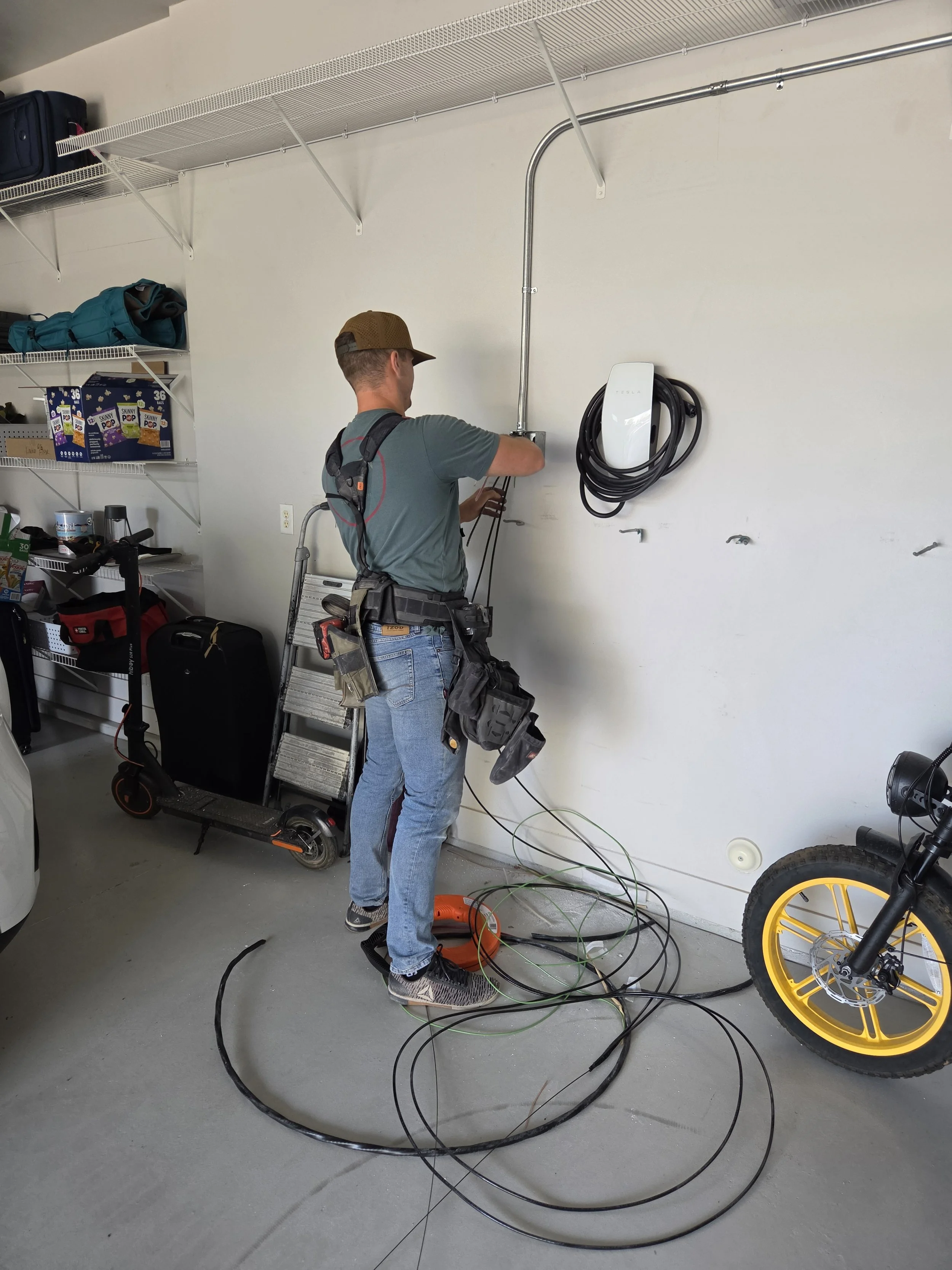 A man in a gray T-shirt, jeans, and a brown cap is working on an electrical outlet or cable on a white wall in a garage. He is standing on a toolbox, surrounded by cables and tools, with shelves of supplies and a scooter nearby, and a Tesla Electric vehicle charger mounted on the wall.
