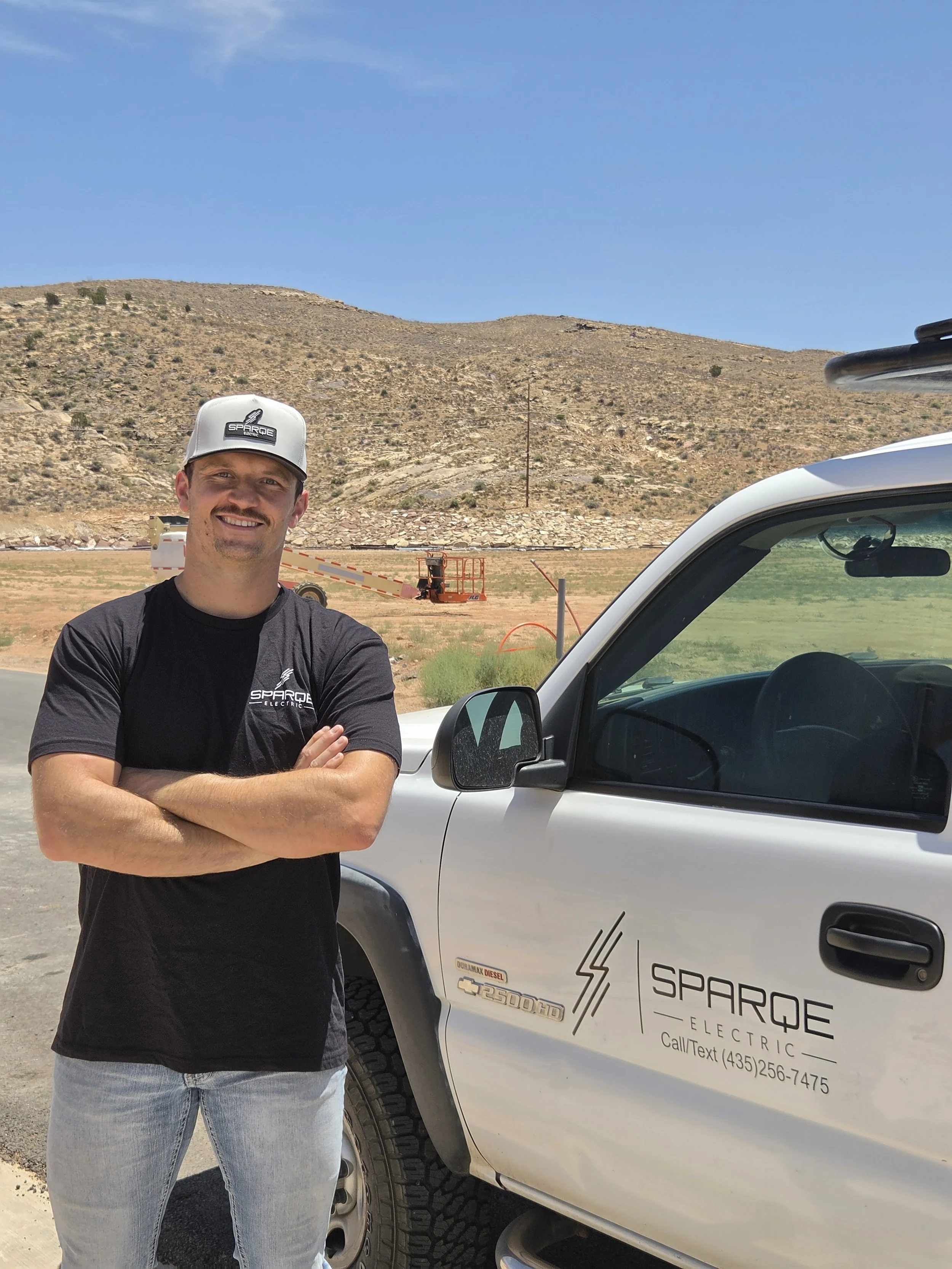 A man with crossed arms standing next to a white vehicle with company logo and contact information, smiling outdoors with a hilly, dry landscape and blue sky in the background.