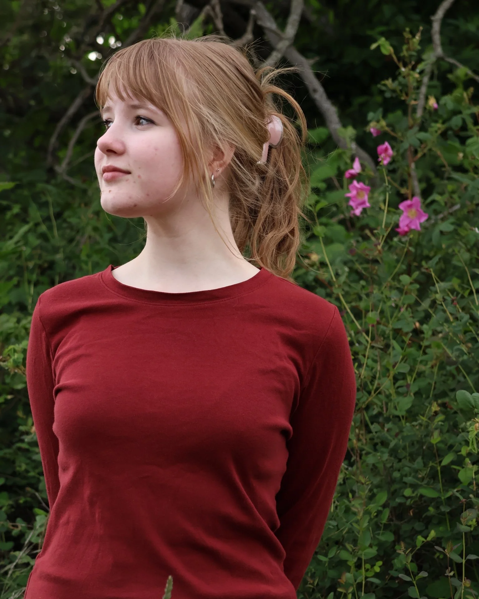 A young woman with red hair and light skin standing outdoors in front of green foliage and pink flowers, wearing a red long-sleeved top.