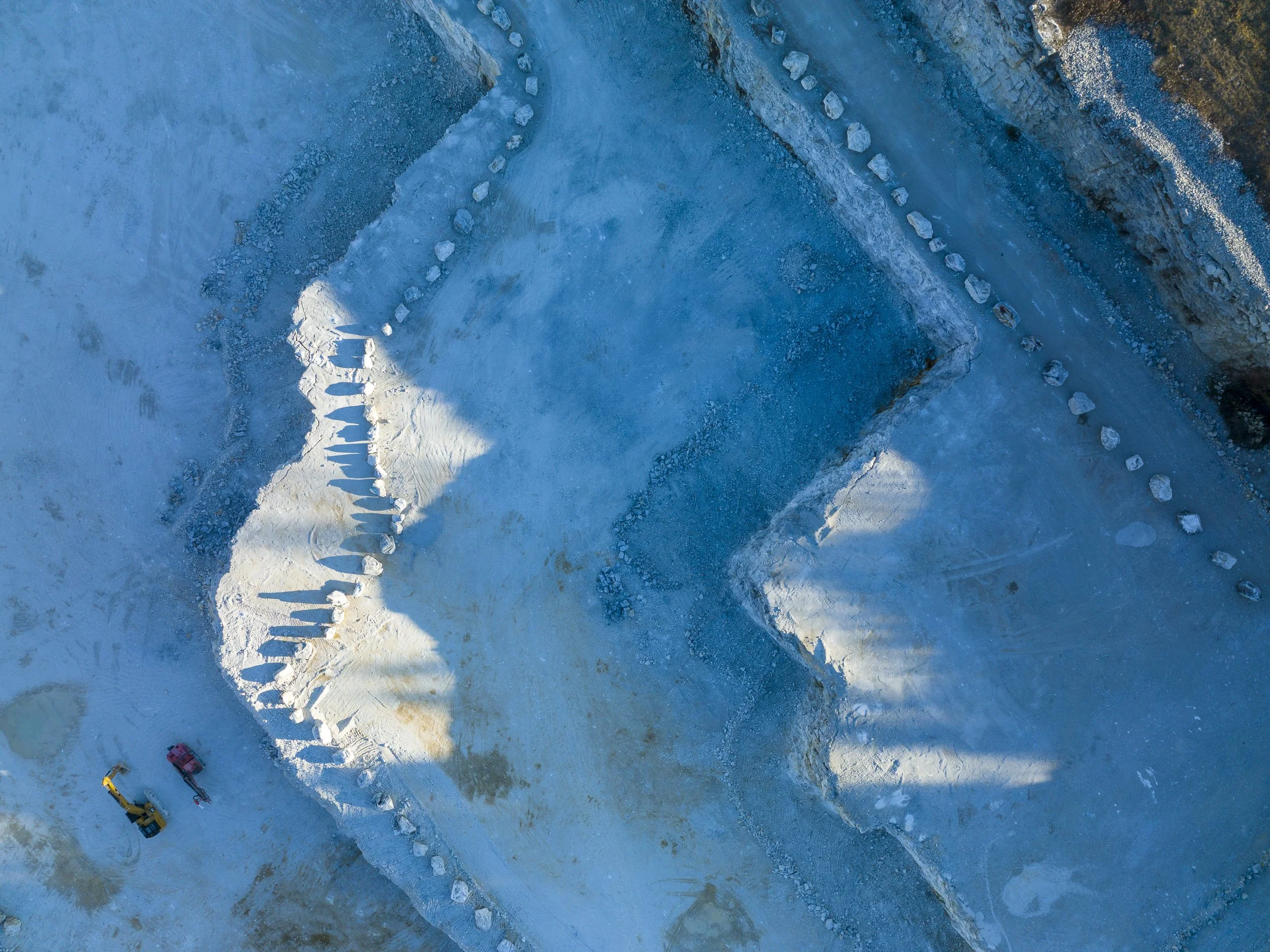 An aerial view of snow-covered ground with a line of three construction vehicles—an excavator, a dump truck, and a roller—parked on the snow, with vehicle tracks leading away.