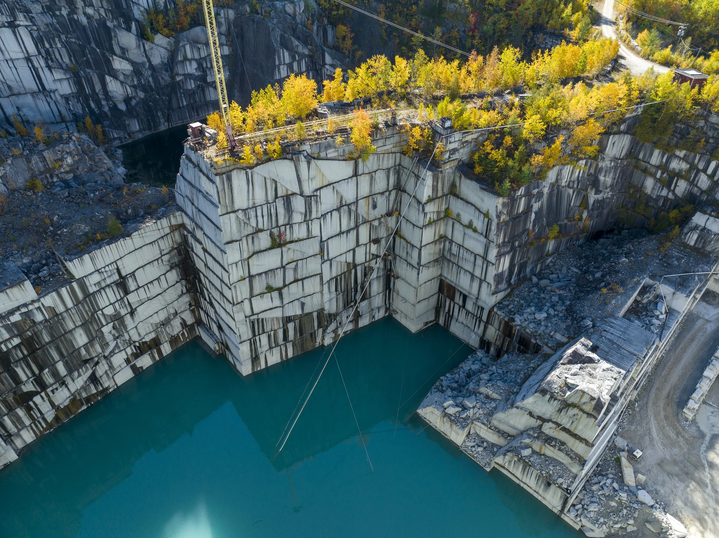 Aerial view photo of a large quarry with white stone walls, yellow trees, and a body of turquoise water at the bottom.