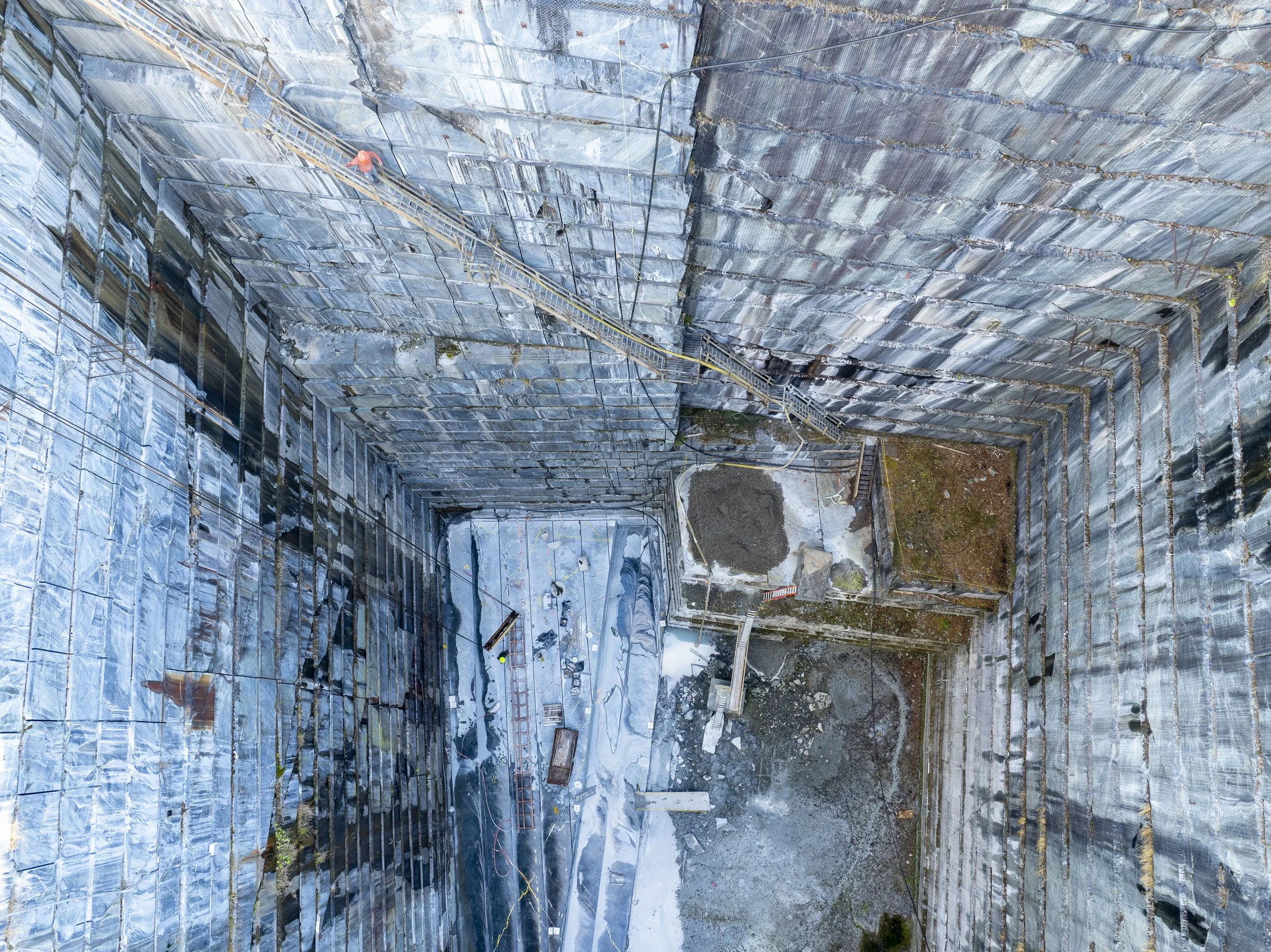 Aerial view photo of a large, deep quarry with steep, rocky walls. The bottom is covered with gravel and some construction materials. There are ladders and safety ropes along the walls for workers.