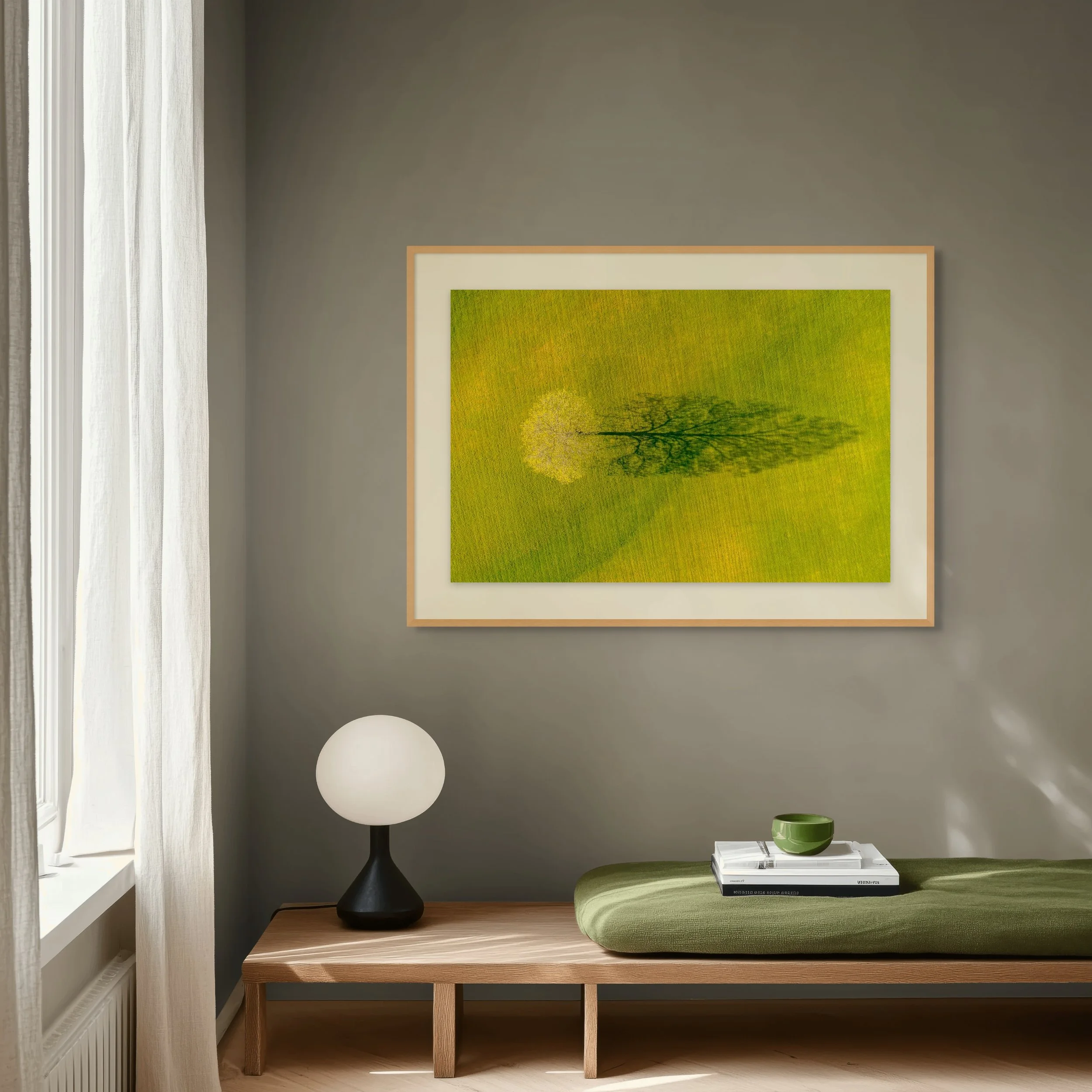 A minimalist interior with a wooden table holding a black lamp and a green bowl, a green cushioned seat, and a framed photo of a tree on a gray wall, with sunlight coming through a window with white curtains.