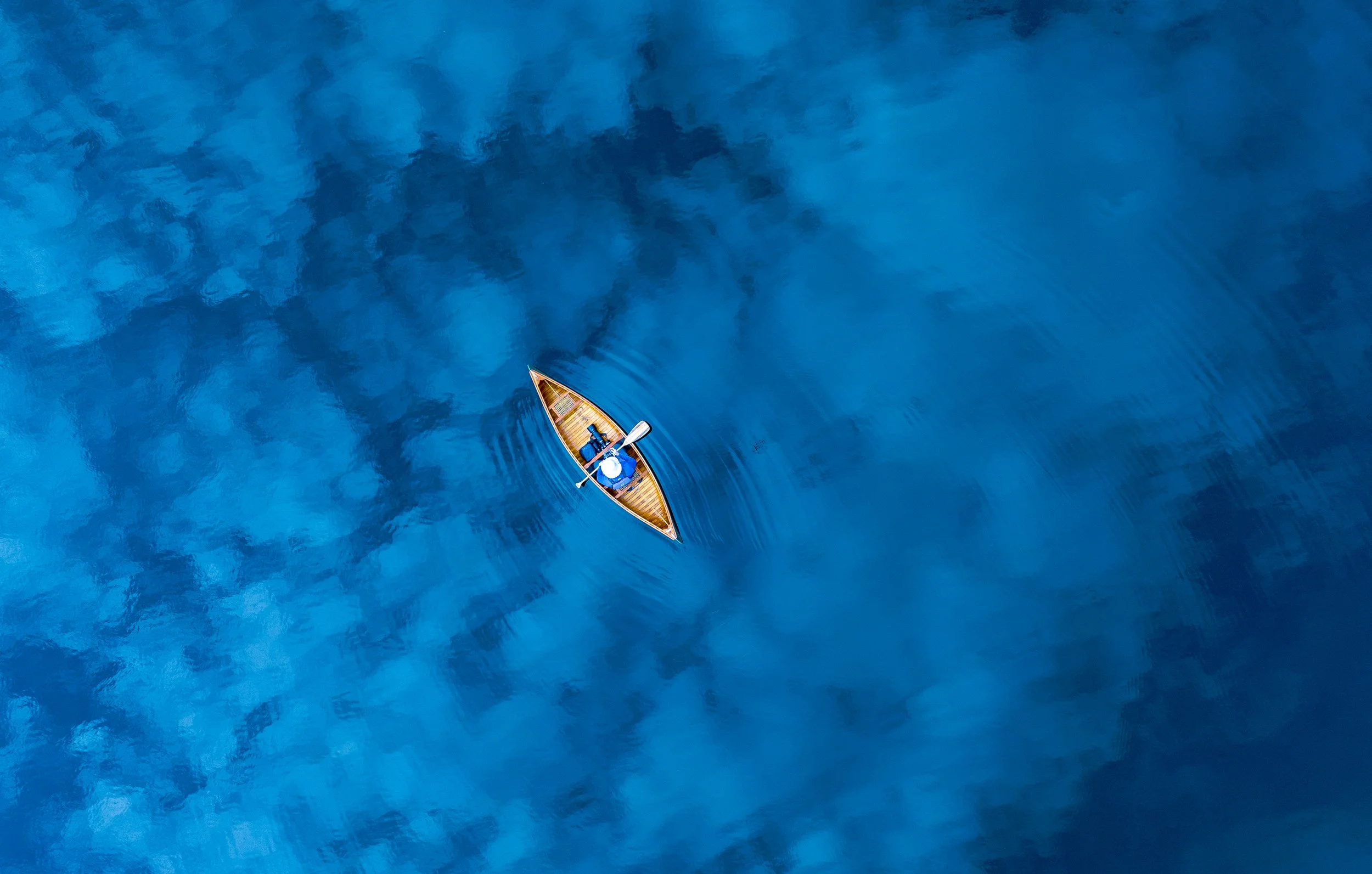 A person rowing a small wooden boat on calm, deep blue water, viewed from above through aerial photography.