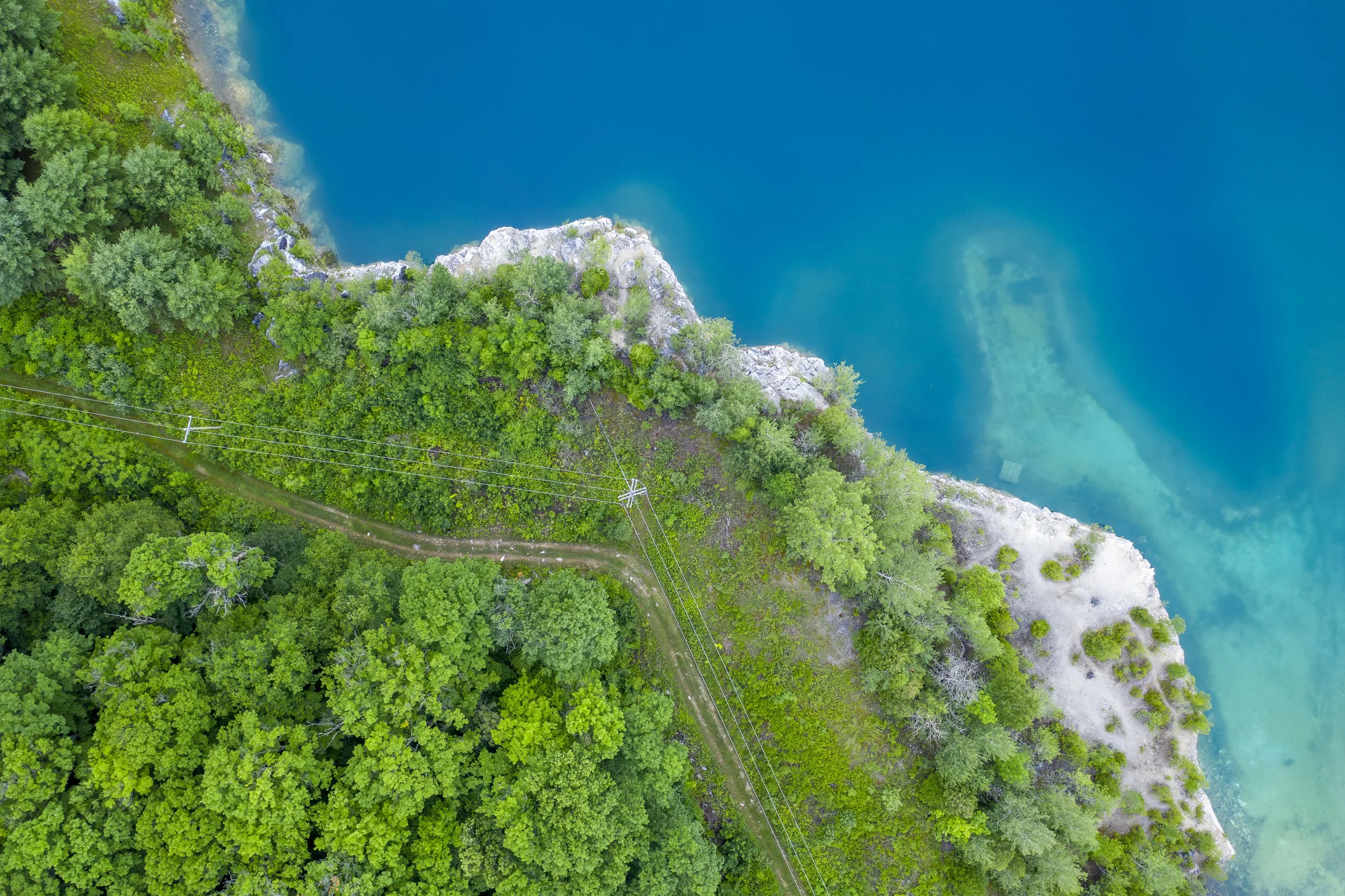 An aerial view landscape photo of a lush forest along the edge of a clear blue body of water, with a small grassy path running through the trees and power lines crossing the forest.