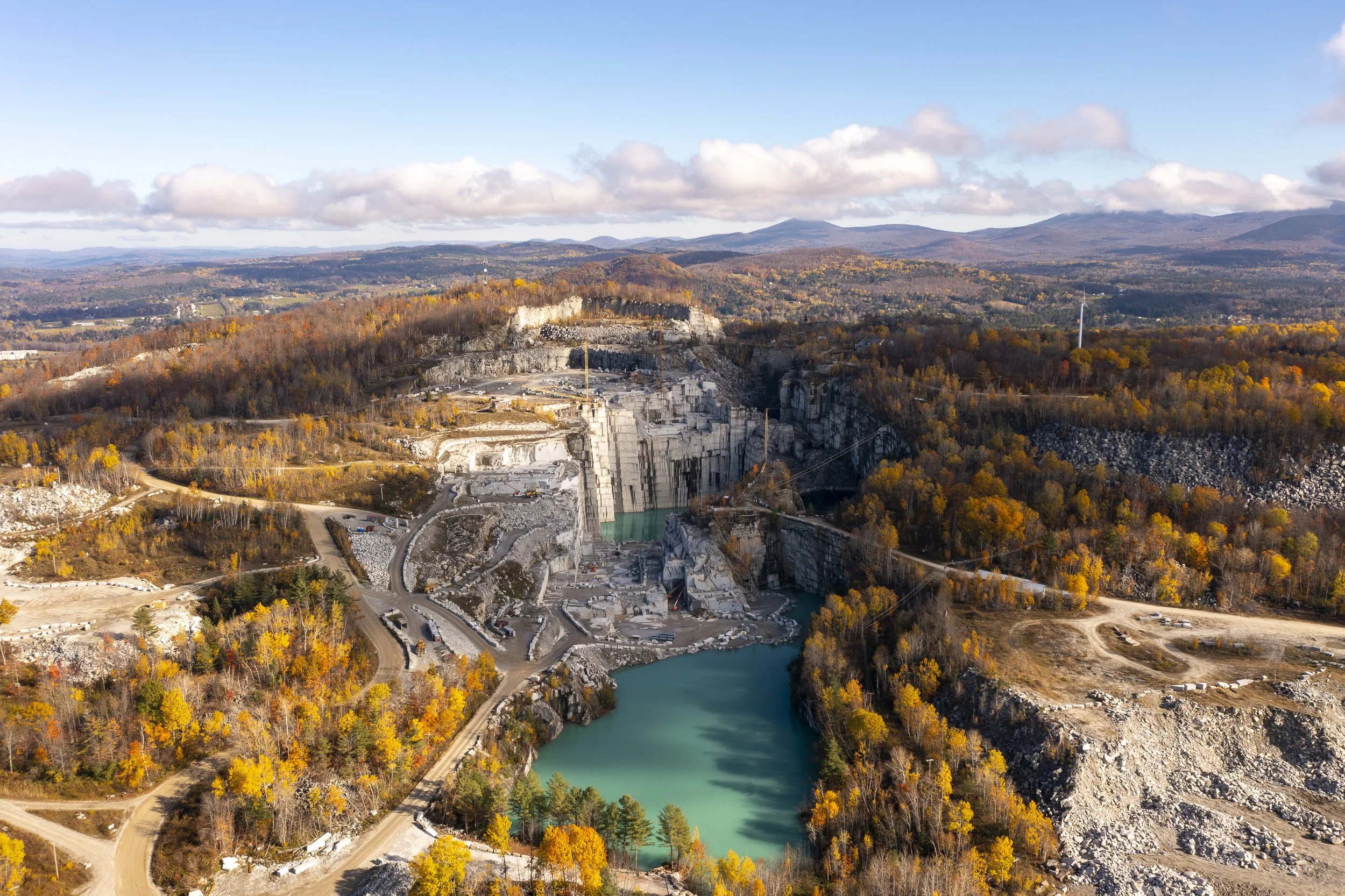 Aerial drone photo of a quarry in a mountainous region during fall with trees in shades of orange and yellow, and a river flowing through the landscape.