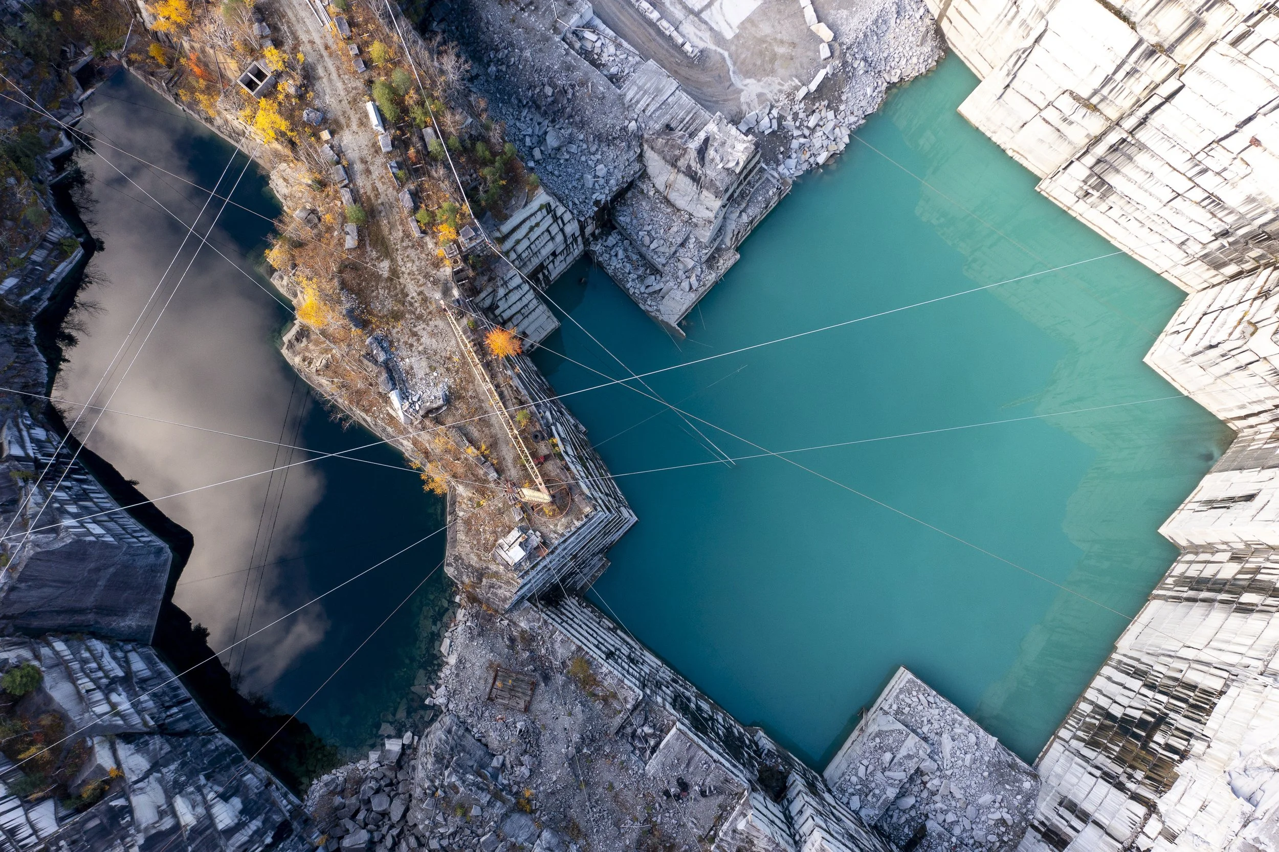 An aerial view of a large quarry with turquoise water, surrounded by rocky cliffs and some autumn-colored trees.