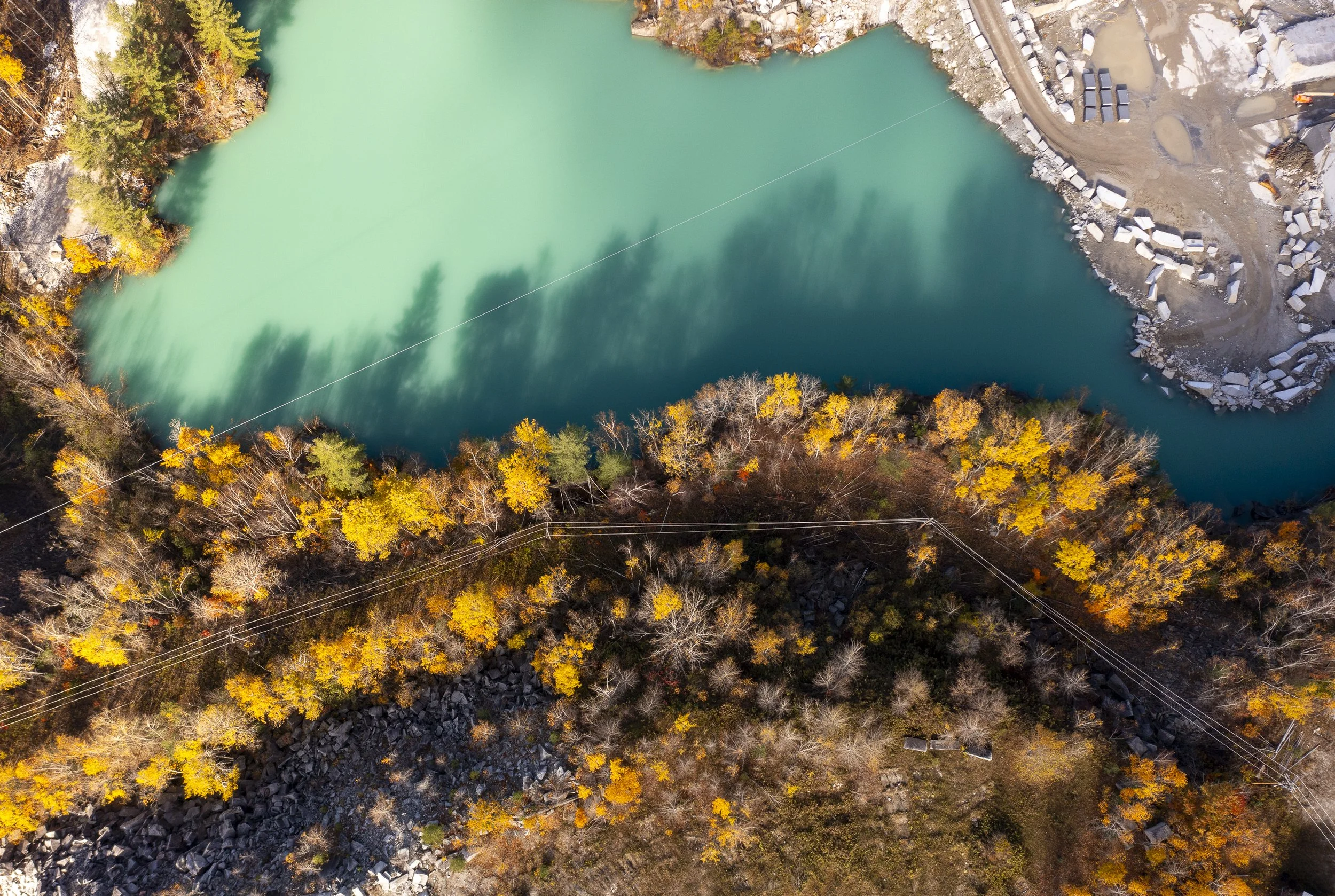 An aerial landscape photo of a turquoise lake surrounded by trees with autumn foliage, rocky shoreline, and construction site.