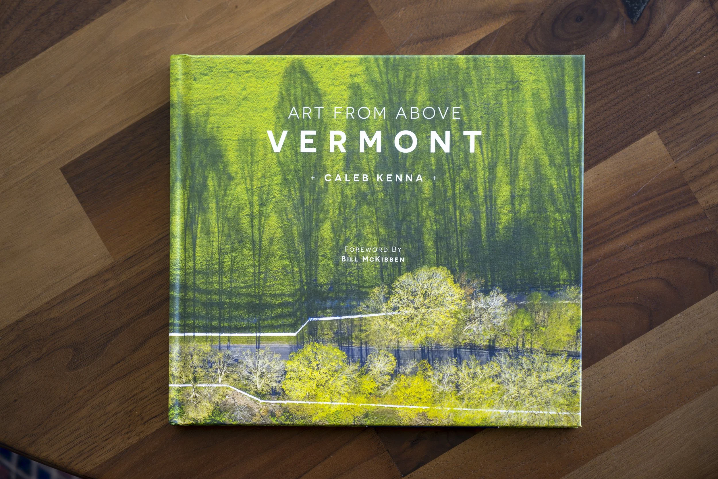 A book titled 'Art from Above Vermont' by Caleb Kenna resting on a wooden surface, featuring a cover image of a forest with trees.