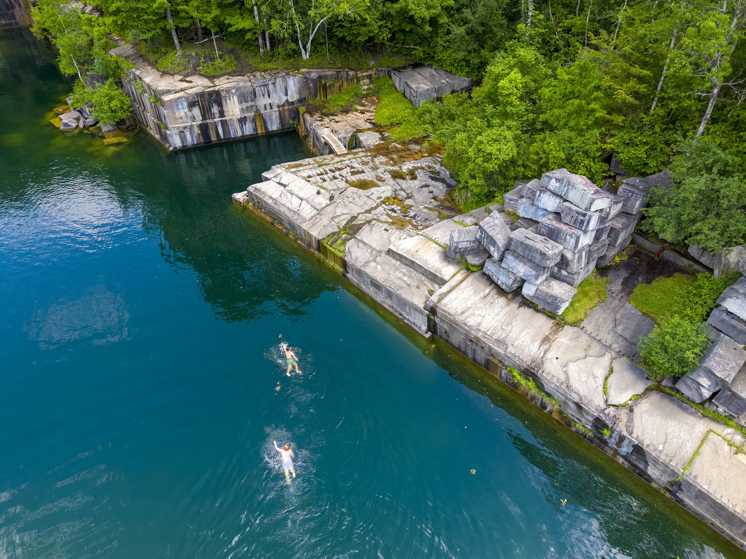 Aerial drone photo of a dam with water flowing over concrete steps surrounded by green trees and a rocky shoreline. 
