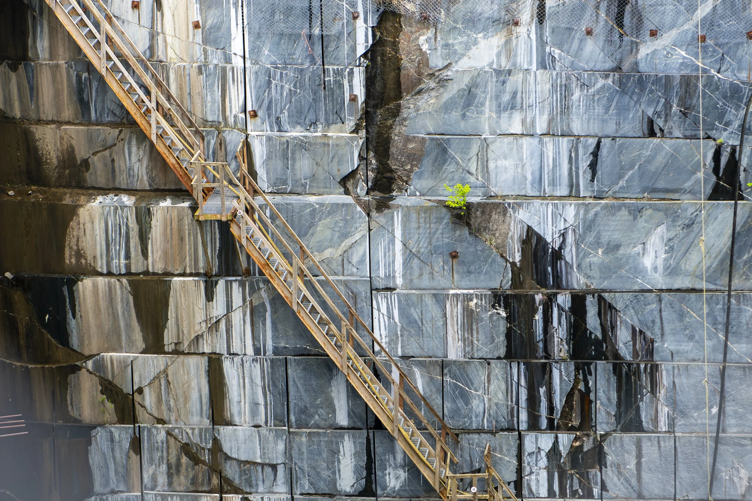 A photo of a large rock quarry with a large vertical rock face and a rusty metal staircase built into the side for access, with small green plants growing in cracks.