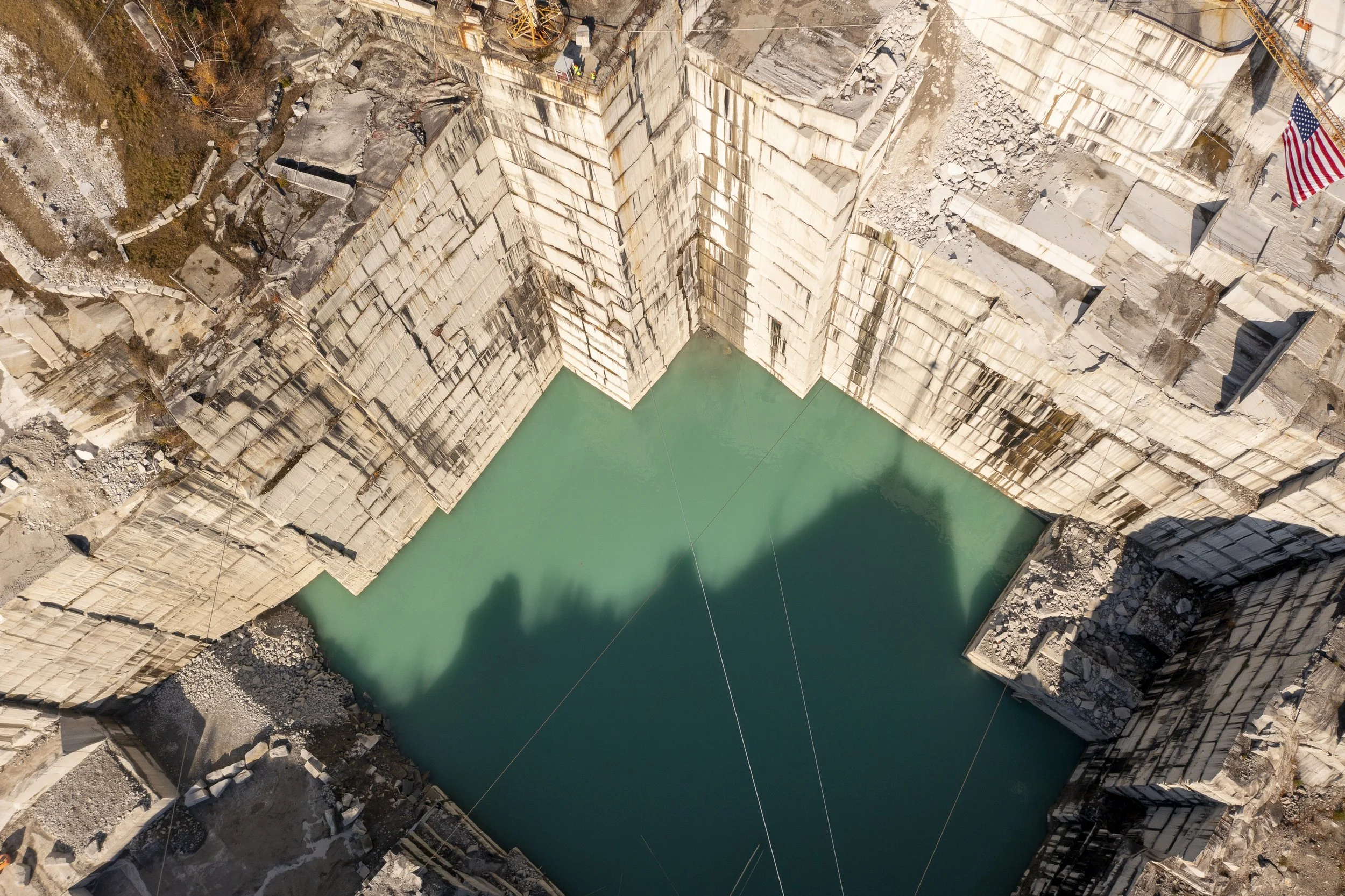 Aerial drone photo of a quarry with high concrete walls surrounding a turquoise water reservoir, with construction workers on top and an American flag on the right.
