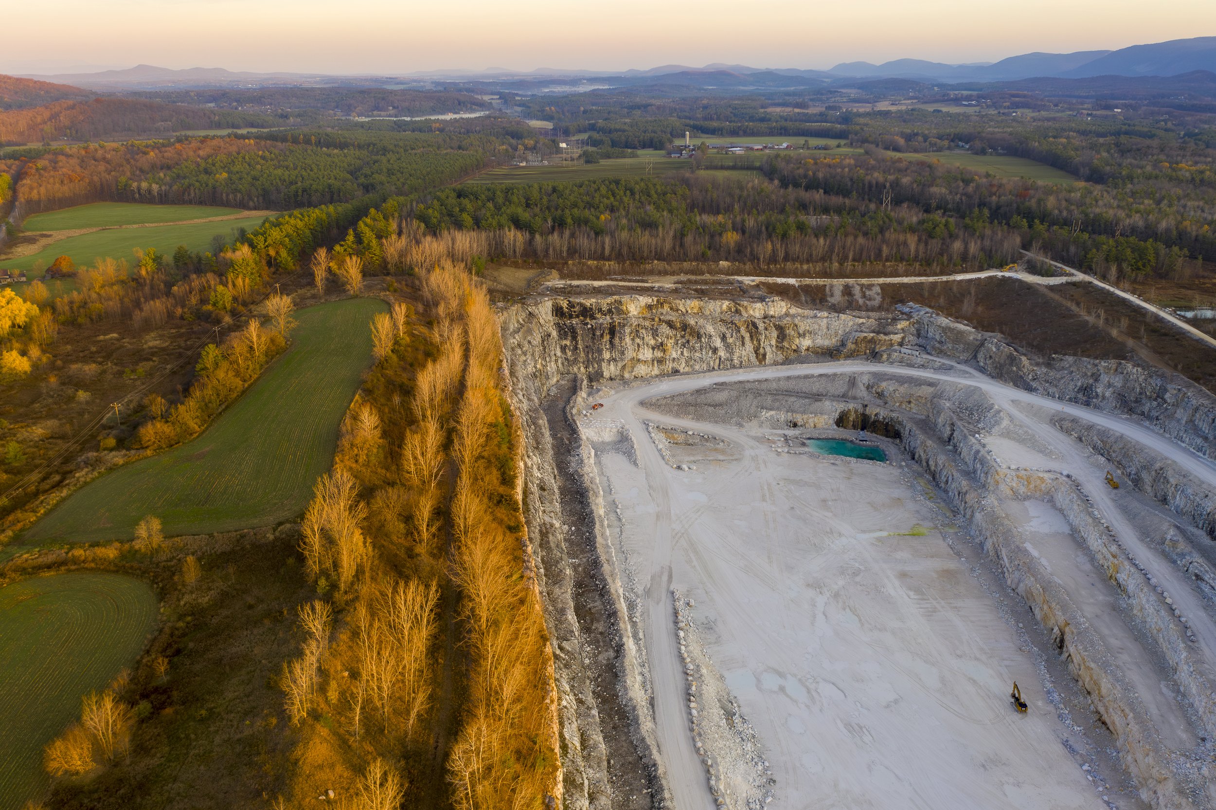 An aerial photo of a landscape showing a quarry partially dug into the hillside with construction equipment, surrounded by trees with fall foliage, green fields, and distant mountains at sunset.