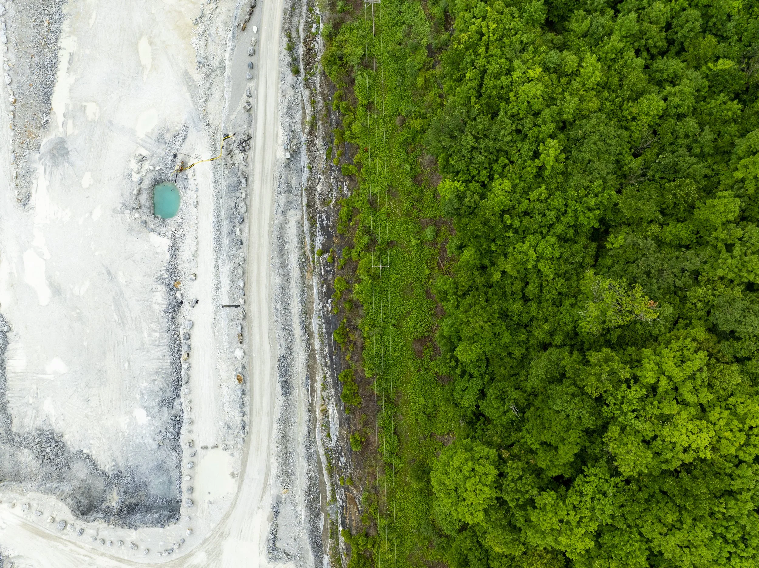 Aerial drone photography of a quarry with a small body of water, rocky terrain, and a dirt road, contrasted with a dense green forest on the right.