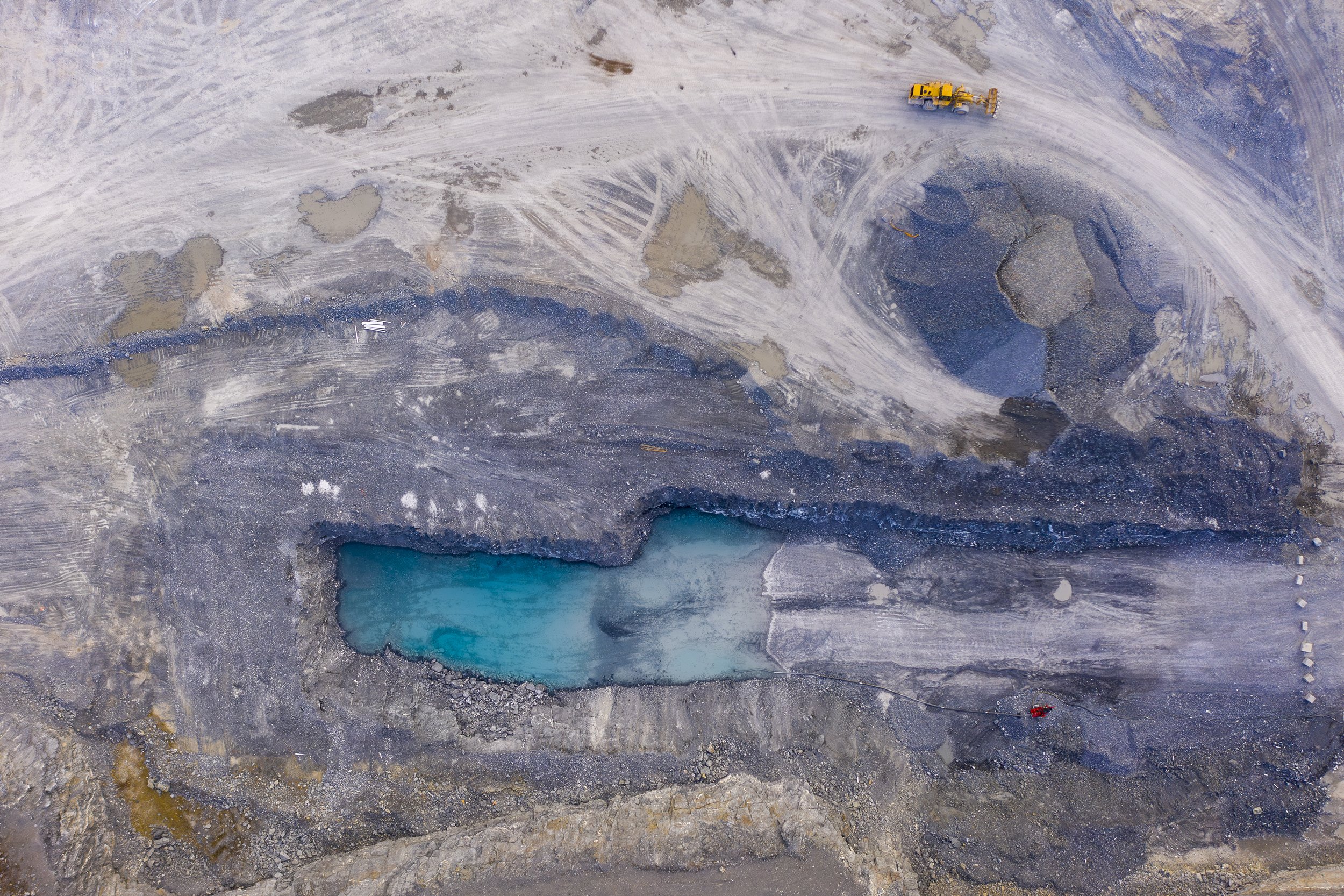 An aerial view photo of a large excavation site with a visible blue-green lake inside a depression, surrounded by gray earth and scattered construction equipment.