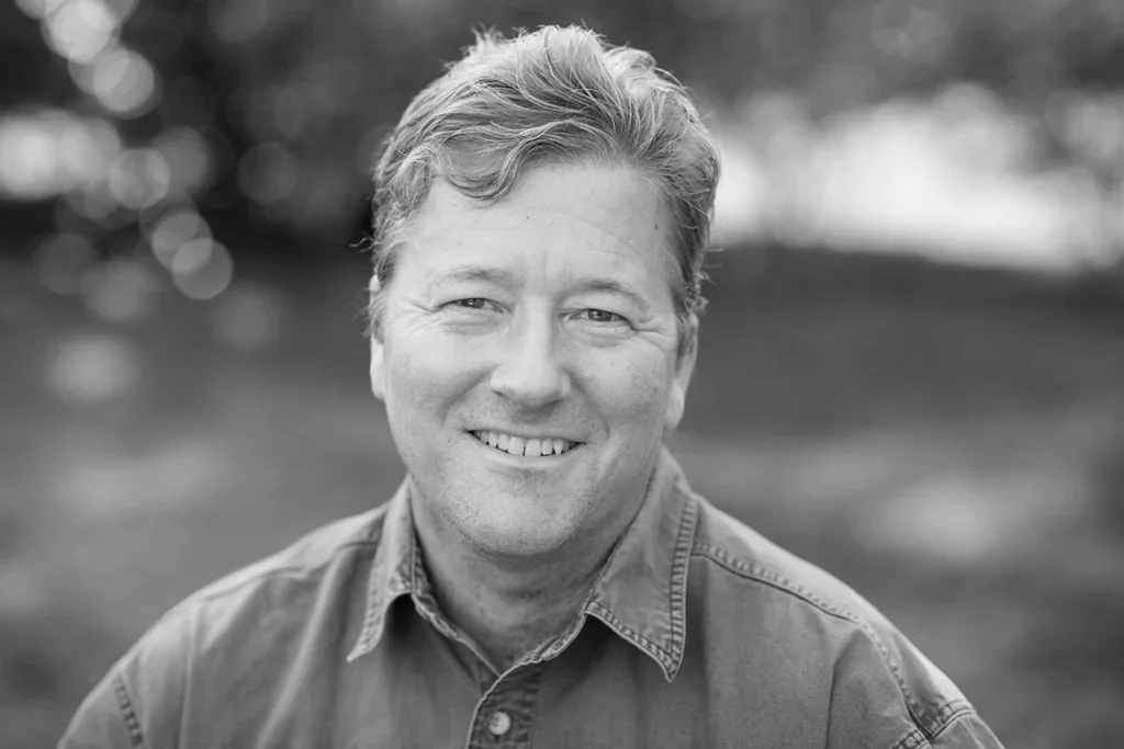 A smiling middle-aged man with wavy hair, wearing a collared shirt, outdoors with blurred greenery in the background.