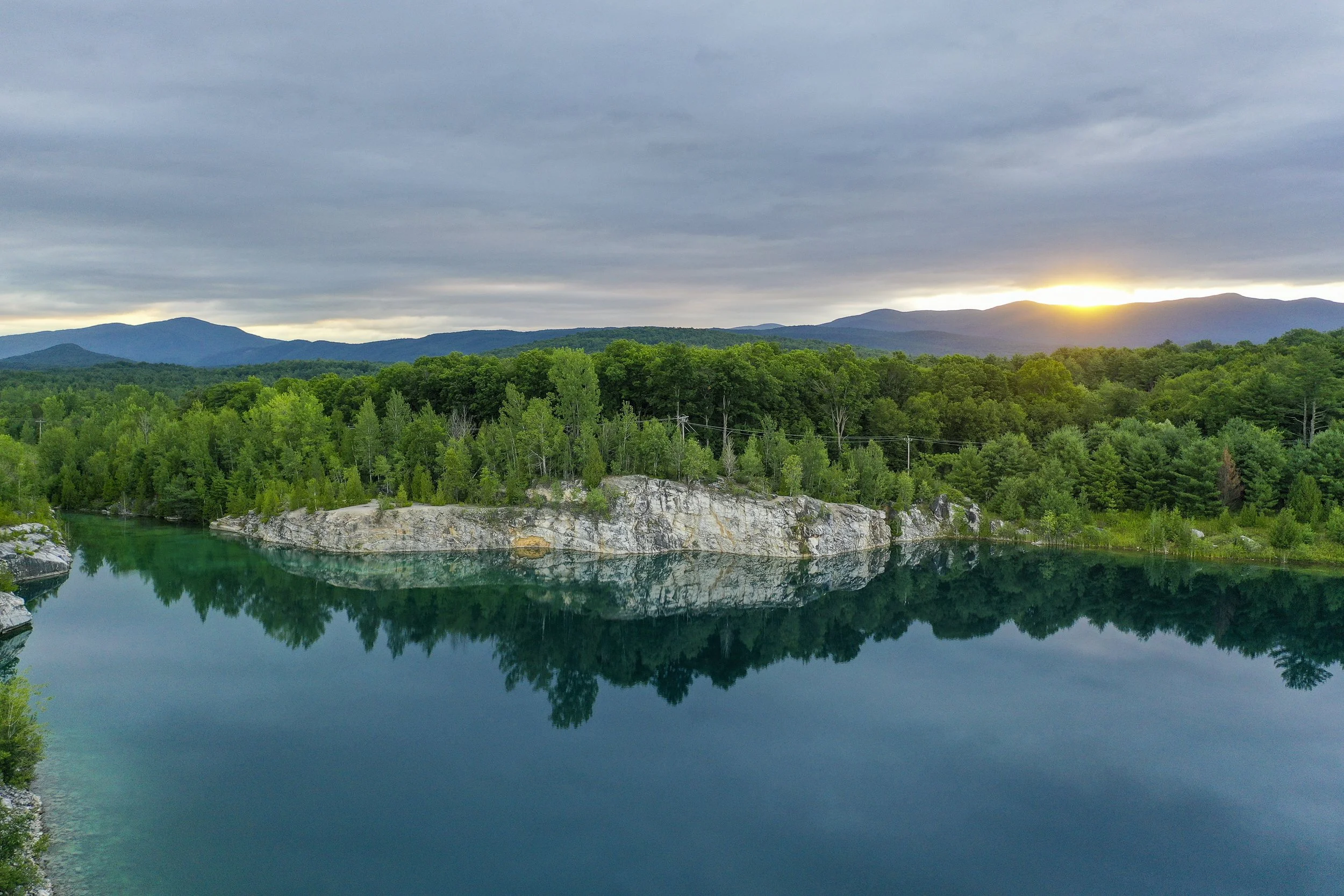 Aerial landscape photo of a serene landscape of a calm river reflecting green trees and rocky shores, with a dense forest, rolling hills, and mountain range in the distance under a cloudy sky during sunset in Vermont.