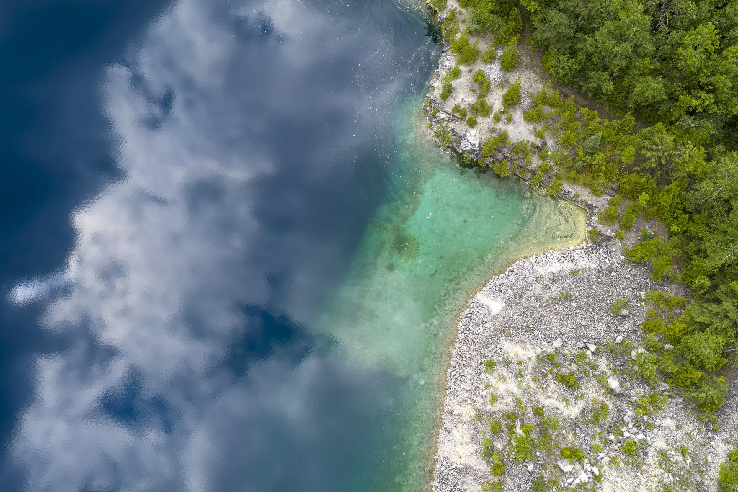 Aerial drone photography of a body of water with shades of blue and green, bordered by a rocky shoreline and dense green forest.