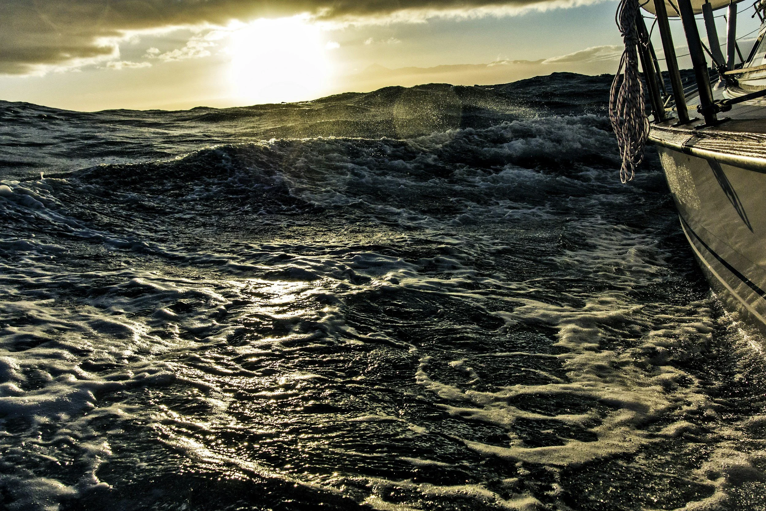 A boat sailing on open ocean waters during sunset with the sun low on the horizon, creating a reflective glow on the water, and a mountain range in the distance.
