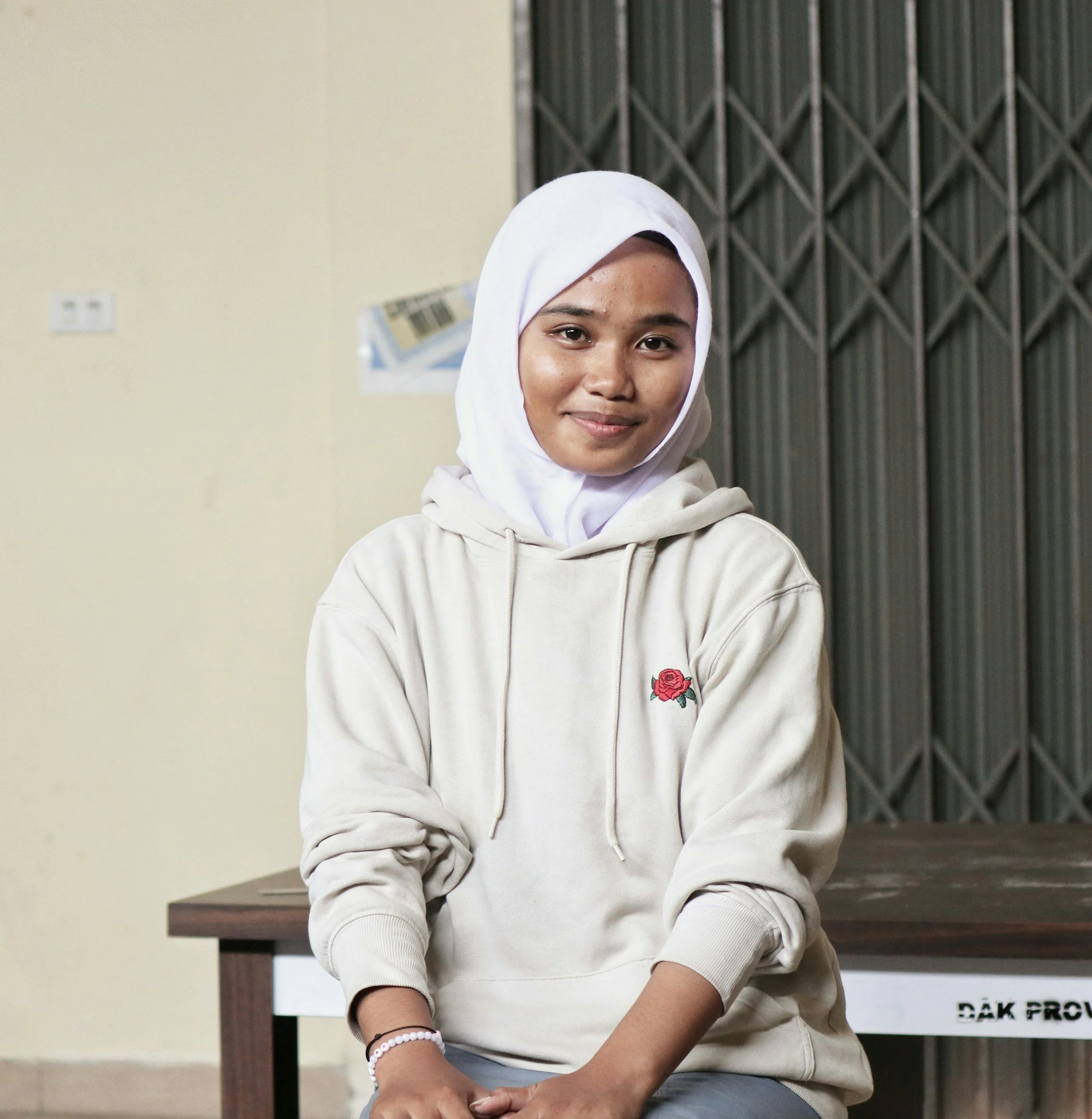 A young woman wearing a white hijab and beige hoodie, sitting at a table with a dark wooden surface, smiling at the camera. Behind her is a beige wall and a black metal gate or door.