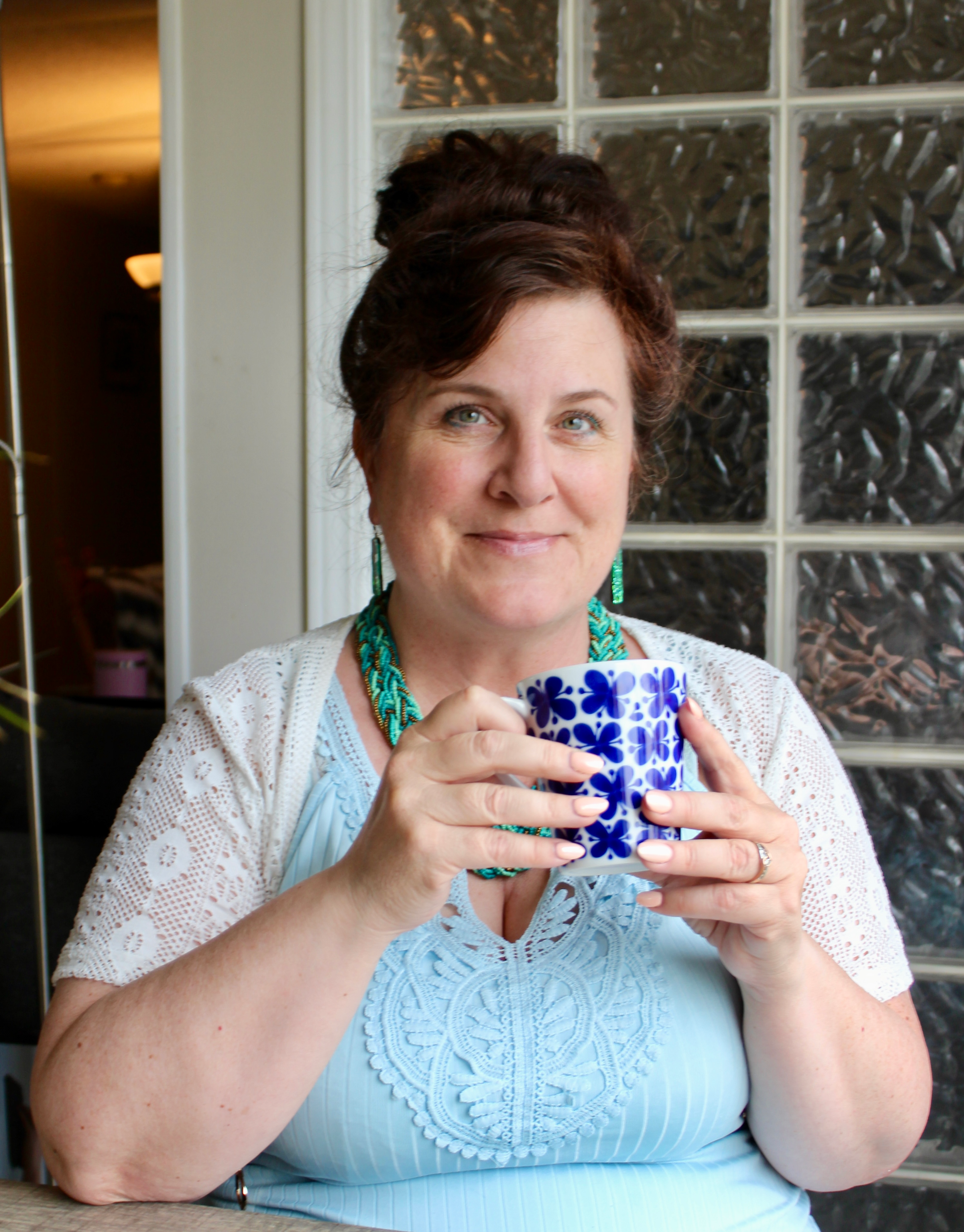 A woman with short dark hair styled in an updo, wearing a light blue top with lace details, turquoise jewelry, and earrings, holding a blue and white floral mug. She is smiling and standing in front of a glass block wall.