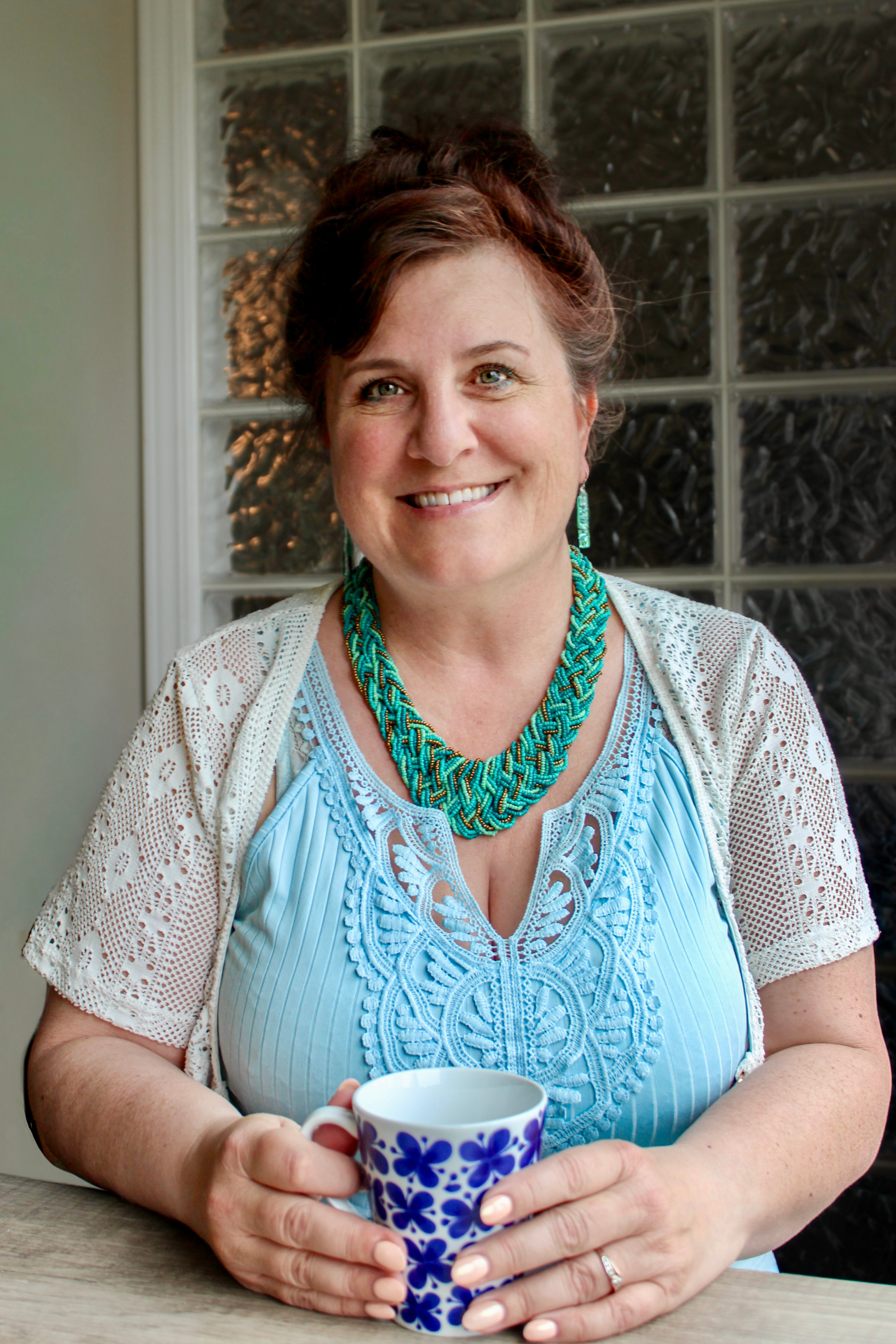 A woman with short, wavy brown hair, wearing a turquoise necklace and matching earrings, sits at a wooden table holding a blue and white patterned mug and smiling while looking at the camera.