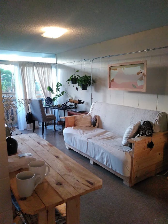 Living room with a white sofa, a wooden table with two mugs, a window with curtains, a desk with a chair, plants, and wall art.