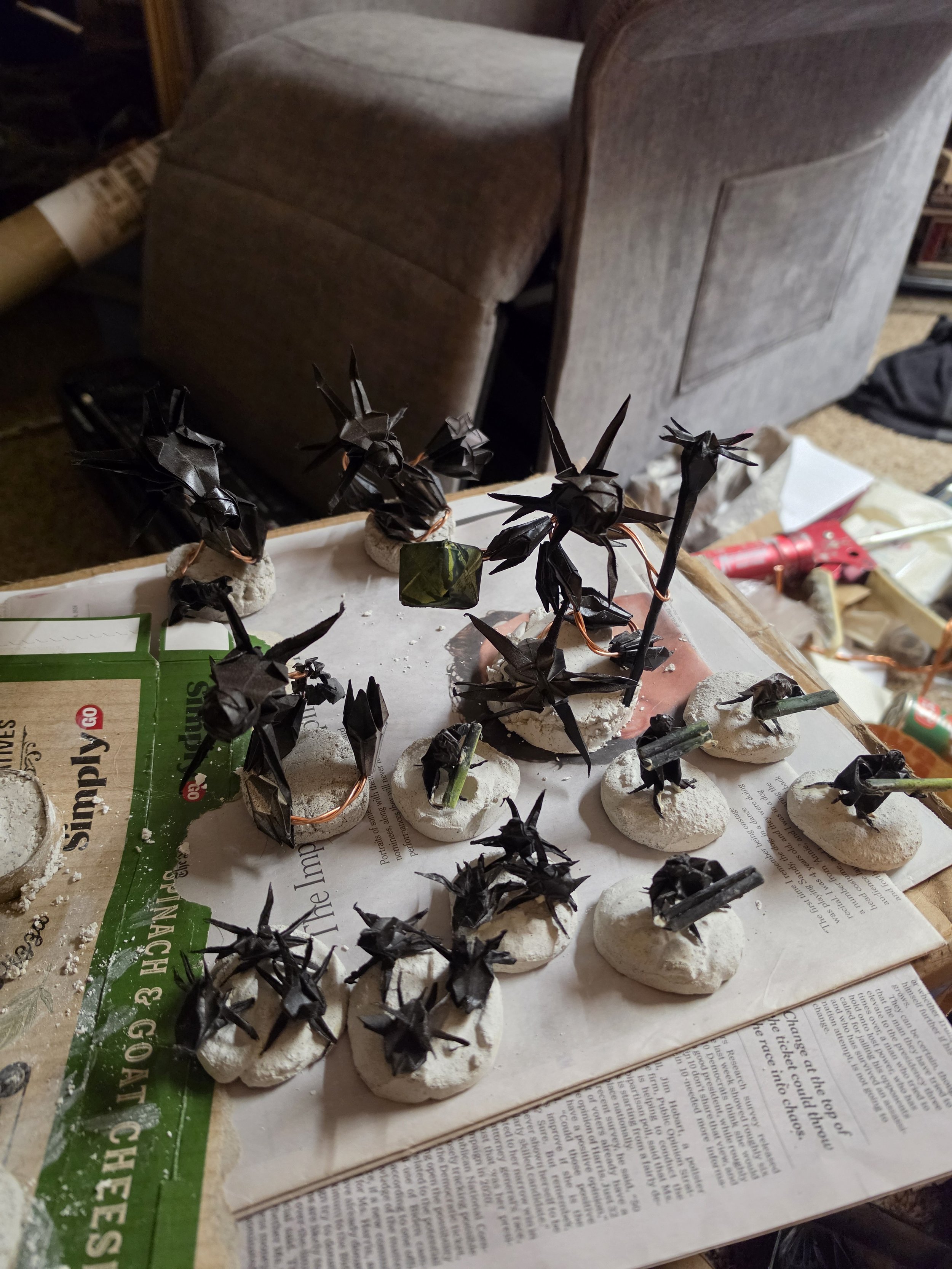 Many small origami black bats perched on white modeling clay bases, arranged on a table covered with newspaper, with a brown upholstered chair and various items in the background.