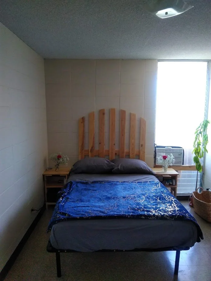 A small bedroom with a bed featuring a wooden headboard, gray pillows, and a blue blanket that appears stained or wet. On either side of the bed are small nightstands, each with a small vase with red flowers. A window with bright light and an air conditioner is seen behind one nightstand. A potted plant and a woven basket are placed on the right side of the room.