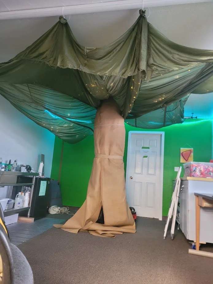 Room with a green wall, a white door, and a makeshift tree made from fabric and a large brown sack, decorated with string lights.